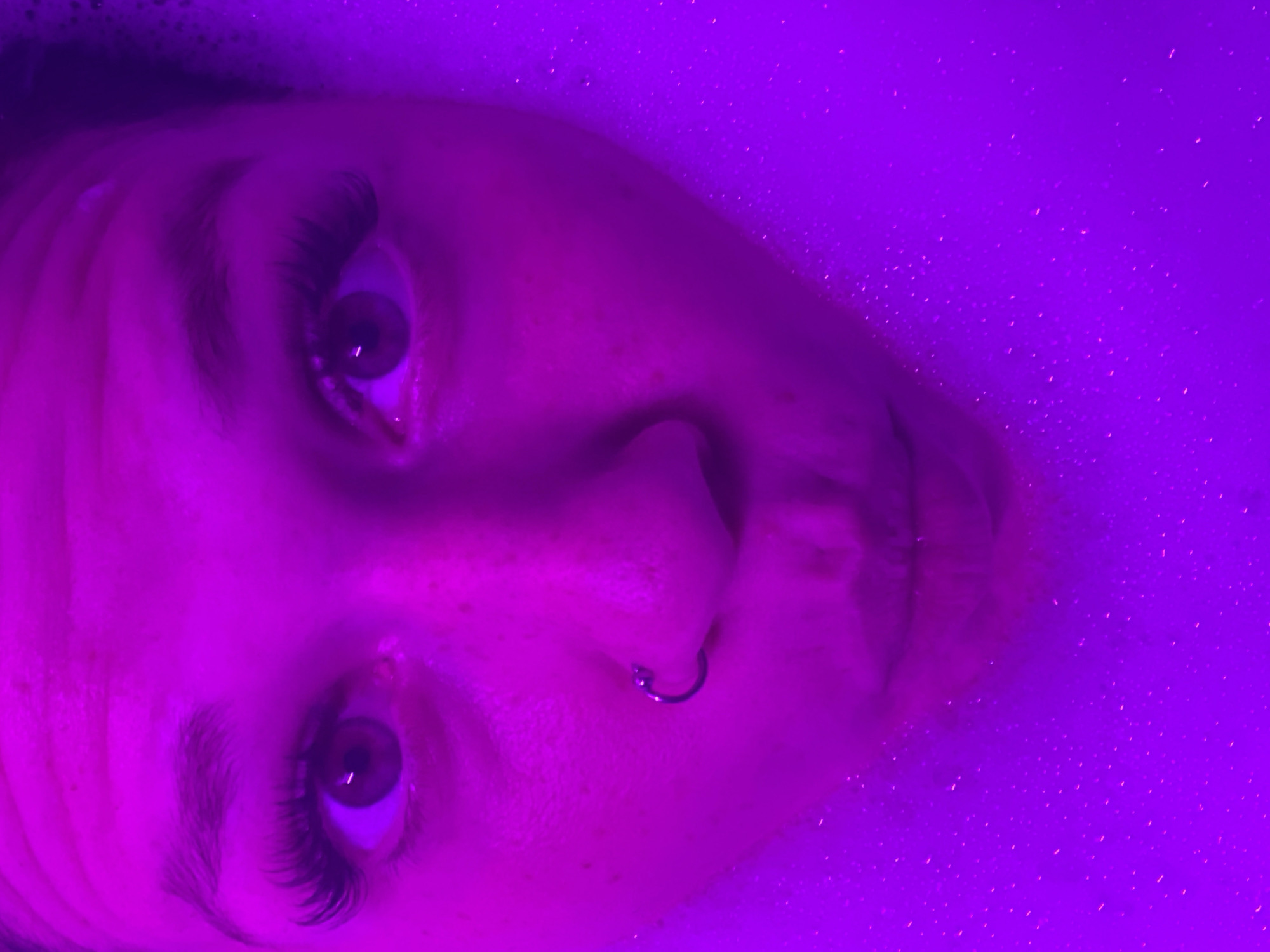 The image is a close-up, overhead shot of a person's face submerged in a bathtub filled with purple-tinted water and bubbles. The person has large, dark eyes with prominent false eyelashes and a nose ring. Their lips are pursed. The lighting is a vibrant magenta hue, creating an atmospheric and slightly surreal effect. The bubbles are visible around the person's face, indicating they are in a bath. There is no information in the image to suggest a location in Chaikovsky, Russia.