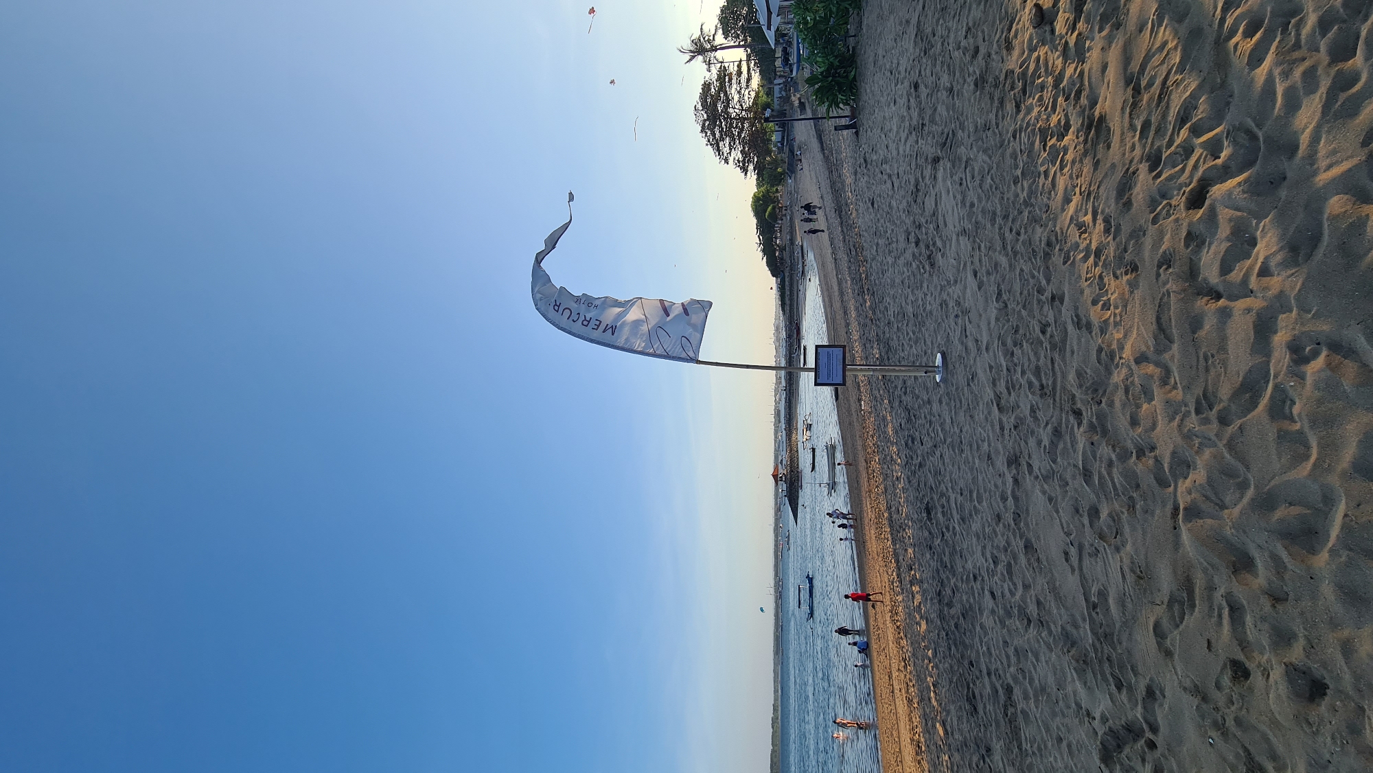 This media file depicts a beach scene in Denpasar, Indonesia, likely during the late afternoon or early evening as indicated by the warm, soft light casting long shadows on the sand. The image is oriented vertically.
The primary subjects are a sandy beach stretching along the coastline and the calm ocean water. Scattered on the beach are a few people, some walking along the shore, while others are closer to the water. Several small boats are visible on the sea, suggesting a leisurely coastal setting. A prominent object is a flag on a pole, fluttering in the breeze. The flag is white with dark lettering. Visible text on the flag reads "MERCUR HOTEL". Next to the flag pole, a small information board or sign is also present. In the background, trees and some buildings can be seen, hinting at the proximity of a resort or town. The sky is a clear, bright blue, indicating pleasant weather. The overall mood conveyed is tranquil and relaxed, characteristic of a beach day.