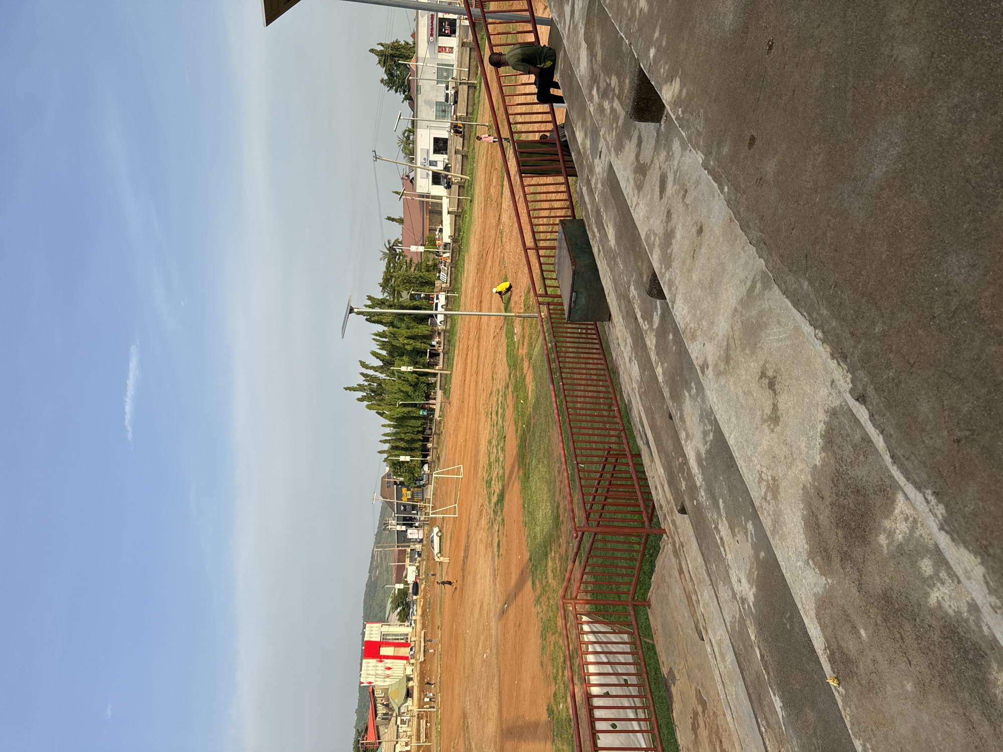 This is a media file that shows a sports field, likely a football pitch, in Abuja, Nigeria. The field is primarily dirt with patches of green grass. There are goalposts visible on the field, indicating a sporting event or practice.
In the foreground, a concrete grandstand with red metal railings is prominent. Several people are visible on the stairs of the grandstand, and one person is standing on the steps, looking towards the field. Another person, wearing a green shirt, is leaning over the railing on the stairs. Further down the stairs, a small yellow object, possibly a toy or a piece of equipment, is resting on the concrete.
In the background, there are buildings, trees, and power lines. The sky is a pale blue, with some faint clouds. The overall lighting suggests it is daytime, likely late afternoon given the angle of the sun and the soft light. The scene appears to be a casual setting, possibly a community sports ground or a school field. No specific text is clearly legible in the image.