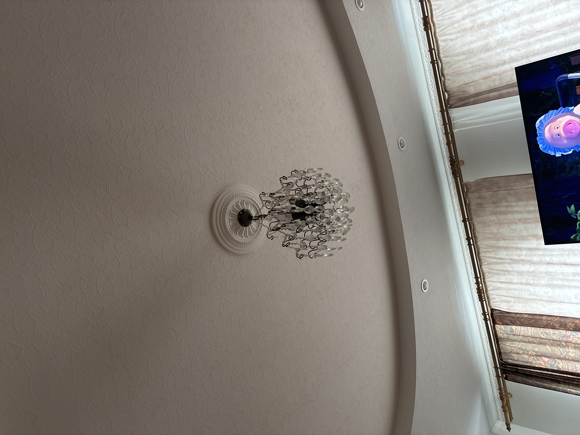 The image shows a view looking up at a ceiling with a decorative chandelier in a room. The ceiling is textured, with a slightly off-white color. The chandelier is ornate, featuring a circular base with decorative elements and multiple strands of clear crystals. The room has a curved architectural detail, including recessed lighting. Part of a television and window are seen in the upper right corner. The window has curtains. There are no people in the image. There is no information indicating the location is in Ferma, Russia.