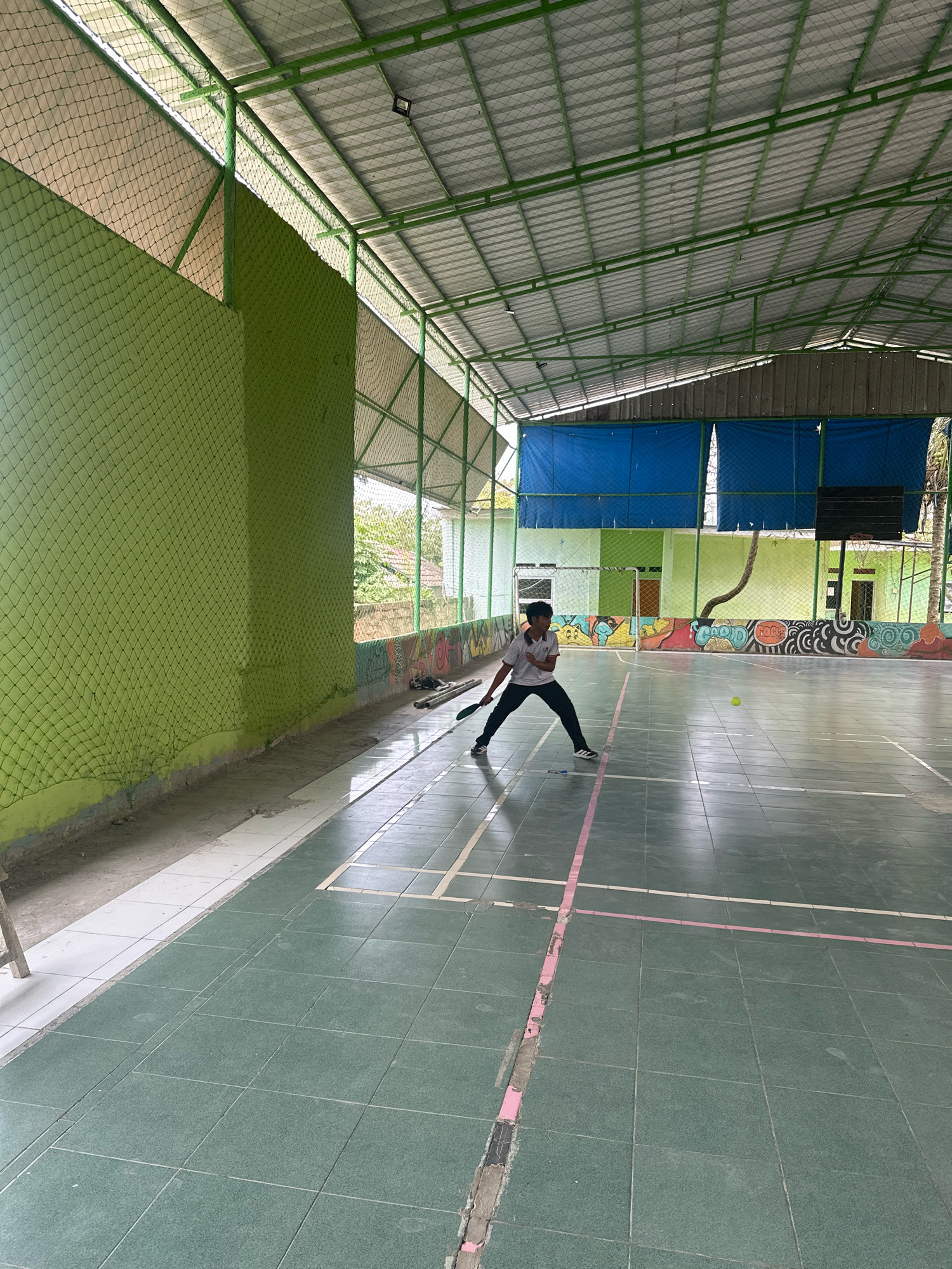 In Saruni, Indonesia, a young male is playing badminton in an indoor court. He is wearing a white polo shirt and black pants and is in the process of hitting a yellow shuttlecock with his racket. The court has a green tiled floor with white and pink court lines. The court is enclosed by green nets and has a metal roof structure. In the background, there are buildings with colorful murals on them and blue tarps covering some areas. The scene suggests a recreational activity taking place in a communal sports facility.