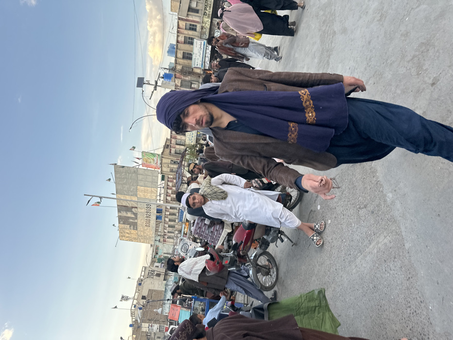 The image depicts a bustling street scene in Quetta City Tehsil, Pakistan, under a clear, light blue sky with hints of scattered clouds suggesting late afternoon or early evening.
In the immediate foreground, a young man walks forward, slightly angled towards the viewer's right. He possesses dark, curly hair and light facial stubble. He is dressed in a dark blue head covering, a dark brown blazer over a dark shirt, and dark blue loose-fitting trousers (shalwar). A long, dark blue scarf with two horizontal gold-patterned bands is draped around his neck. His right hand holds a strand of light-colored prayer beads (tasbih). His expression is neutral, and his gaze is directed slightly downward.
In the mid-ground, to the left of the primary subject, another young man with a neutral expression leans against a red motorcycle. He wears a white shalwar kameez and a white skullcap, with a patterned scarf draped over his left shoulder and open-toed sandals on his feet. The motorcycle's rear rack is laden with red, cylindrical items, possibly merchandise. Adjacent to the motorcycle, a small blue cart or stall displays various items.
The background reveals a busy urban environment with numerous individuals, predominantly men in traditional attire (shalwar kameez, turbans, skullcaps), walking and standing. Several women, dressed in dark, loose clothing, some with headscarves or burqas, are visible on the far right. Multi-story buildings line the street, appearing worn, with various signs in Urdu script. A large billboard in the mid-background prominently displays the partial number "0300 929999". Multiple small flags, some resembling the Afghan flag, are mounted on poles on rooftops and streetlights. The ground is paved, indicating a road or public square. The scene conveys an active, commercial street atmosphere.