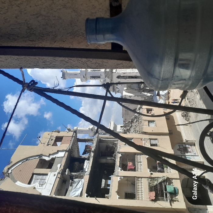 The image depicts a street scene in Khan Yunis, Palestinian Territory, observed from an elevated perspective, likely a window. In the immediate foreground on the right, a large, translucent blue plastic water bottle occupies the lower right quadrant. Dark, thin metal bars or wires, possibly part of a railing or security grille, crisscross the central foreground, partially obscuring the view.
To the left, a multi-story building, likely residential or commercial, exhibits severe structural damage with large sections of its facade missing, exposing interiors and reinforcing bars. Rubble is visible within the building and piled at its base. Clothes, including pink and red items, hang on lines from balconies on the lower visible floors, and a white tarp covers an opening on a middle floor. Street-level signage is partially visible below the damaged building.
A debris-strewn street runs across the midground. Three small figures, appearing to be people, are discernible on the street to the right of the damaged building, facing away from the viewer. In the background, to the right of the street, stands a white mosque with at least two distinct minarets, also showing signs of damage. The sky above is blue with scattered white clouds.