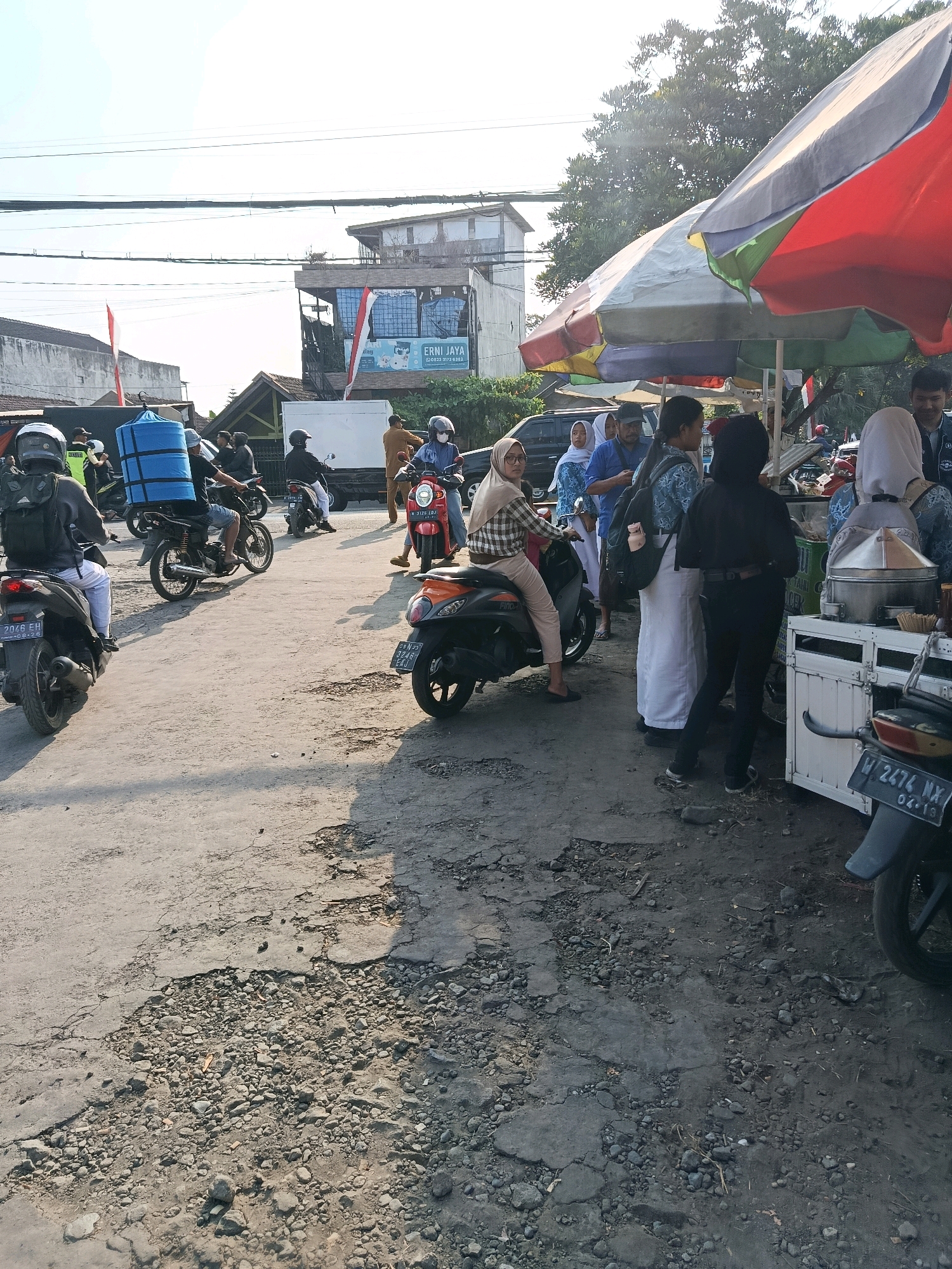This image captures a busy street scene in Bululawang, Indonesia, likely near a market or food stall area. The foreground shows a dilapidated asphalt road with numerous potholes and loose gravel, indicating a less-maintained urban environment. Several motorcycles are present, some with riders, suggesting common local transportation.
In the mid-ground, a group of people are gathered around a mobile food cart or stall, partially covered by a colorful umbrella. Individuals are seen in casual attire and some in what appear to be school uniforms, hinting at a mix of students and local residents. One person is on a scooter, and another is about to mount theirs, indicating ongoing movement and activity. A blue insulated container is visible on the back of one of the motorcycles.
The background features buildings, including one with signage that reads "ERNI JAYA," and trees. Power lines are strung across the sky, typical of many urban and semi-urban settings. The lighting suggests it is daytime, possibly late morning or afternoon, with sunlight casting shadows. The overall atmosphere is one of everyday commerce and local life in a dynamic Indonesian setting.