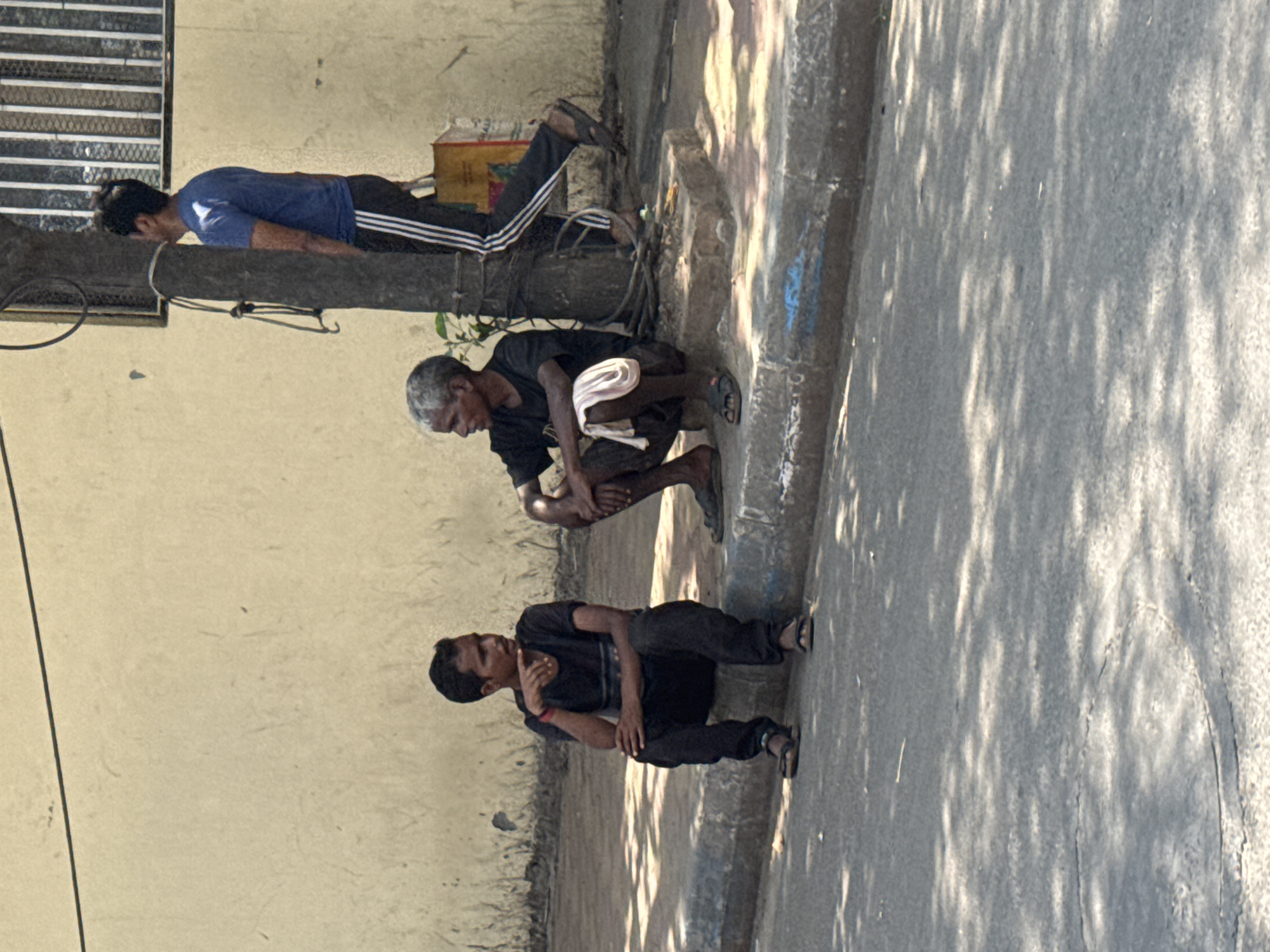 Three men are positioned adjacent to a building and a street in Kolkata, India. The top man, wearing a blue shirt and black pants with three white stripes, is lying prone on a dark, possibly metallic, horizontal structure. Below him, an older man is squatting on the ground, wearing a dark shirt and a light-colored cloth draped over his lap. The third man is also squatting, wearing a dark shirt and pants. The building's wall is light in color, while the street is a darker gray, partially shaded by tree foliage.