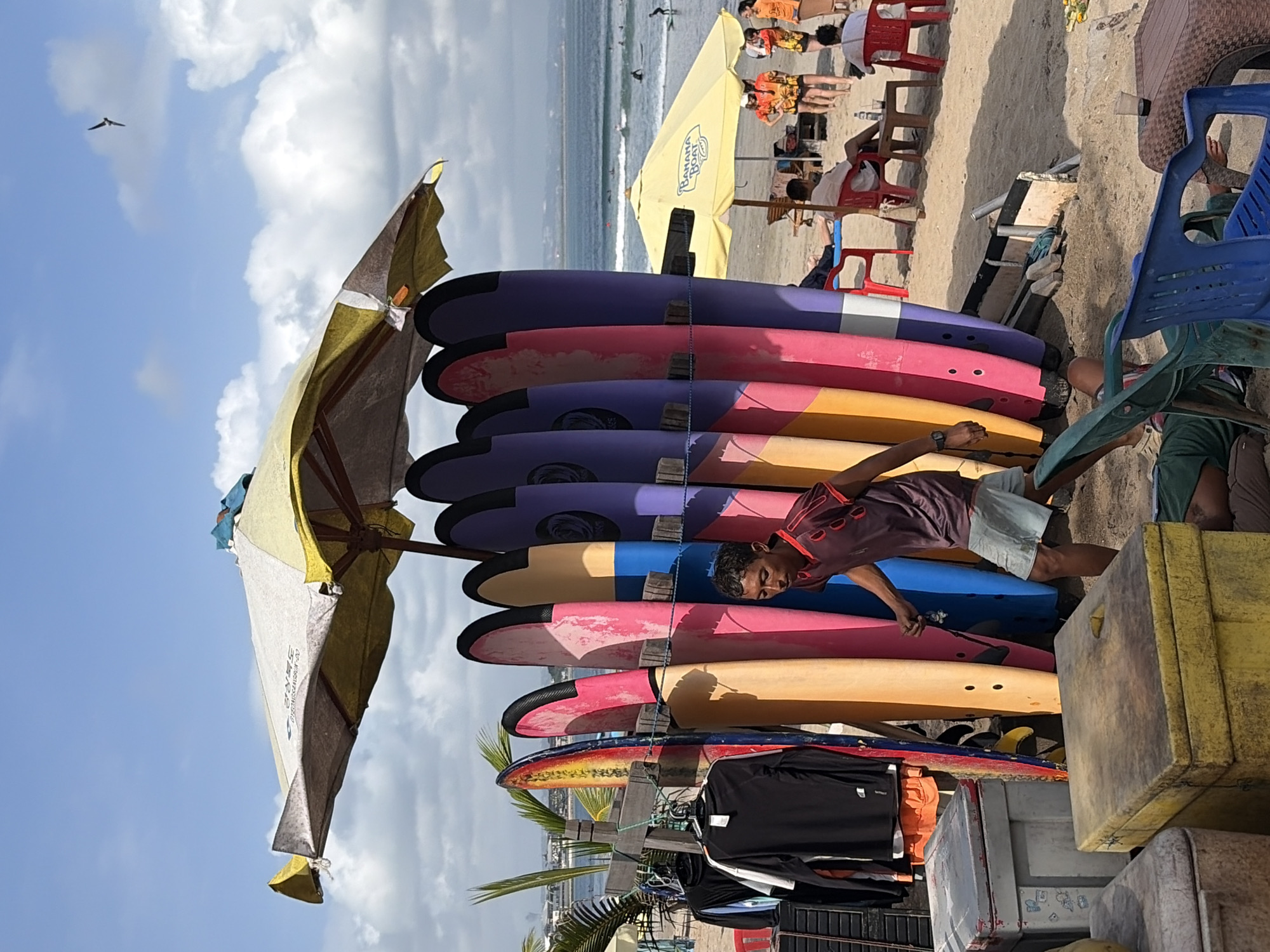 The image depicts a beach scene in Kuta, Indonesia, likely a surf rental area. In the foreground, a young man with dark hair, wearing a maroon t-shirt and light blue shorts, walks to the right. He is positioned near a rack of colorful surfboards, which are lined up vertically. Several surfboards are visible, in shades of yellow, pink, purple, and blue. To the left of the surfboards, several black and white shirts are hanging on a rack. In front of the surfboards, two large, weathered, yellow and gray storage boxes are present. Further back, people are seated on chairs under a yellow umbrella. Beyond the beach, the ocean is visible with several surfers in the water and gentle waves. The sky is blue with scattered clouds, and a bird is flying in the upper right portion of the image. The overall environment is a sunny, outdoor beach setting.