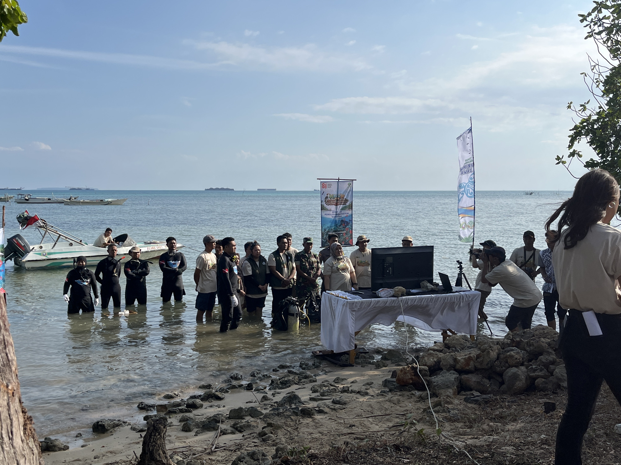 In Kemloko, Indonesia, on a sunny day with a clear blue sky and calm water, a group of people are gathered near the shoreline. The scene takes place in shallow water with a sandy, rocky beach in the foreground.
There are two boats in the water. One is a small motorboat with a Yamaha engine, partially visible on the left, and another appears further out. Several cargo ships are visible in the distance on the horizon.
The main group of individuals are standing in the shallow water. Many are wearing dark diving suits, some with white gloves, suggesting they are divers. Others are in casual or official attire, including t-shirts, shorts, and some in military-style uniforms. There's a table covered with a white cloth, set up near the water's edge, with a laptop and a screen on it. Several people are positioned around this setup, with one person appearing to operate a camera on a tripod. Two large banners are displayed, one with text and images, and another taller, narrower banner with a blue and white design.
The overall atmosphere appears to be an outdoor event or presentation, possibly related to marine activities or environmental conservation, given the presence of divers and banners with Indonesian text. The environment is a coastal area with the ocean stretching out towards the horizon.