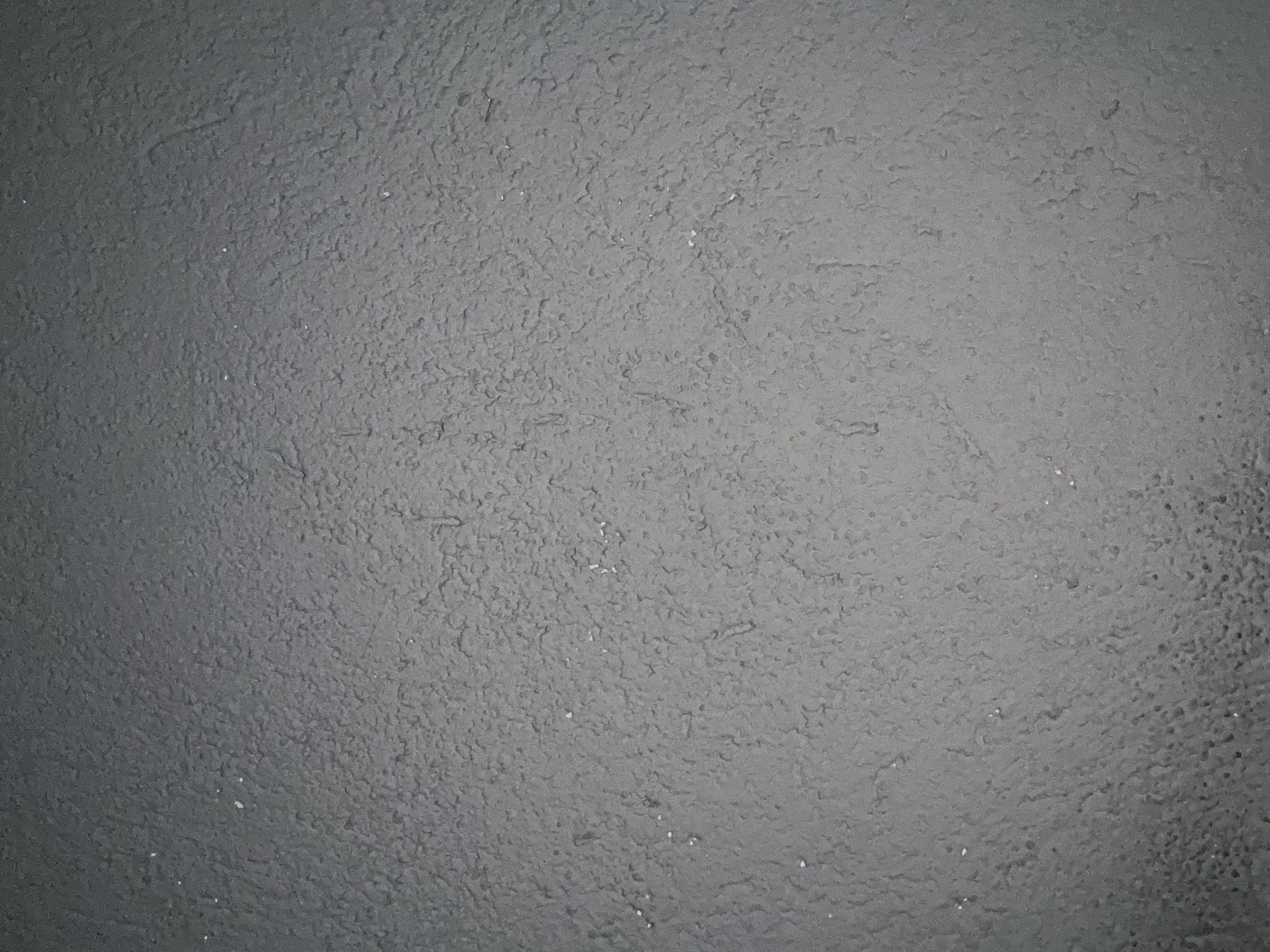 The media file presents a close-up, high-angle view of a dark gray, highly textured surface. The surface is characterized by an irregular, rough, and uneven topography, featuring numerous small bumps, ridges, and depressions. This creates a granular or stucco-like appearance across the entire frame. The dominant color is a consistent shade of dark gray, with subtle variations in lightness and shadow that accentuate the three-dimensional texture. Lighter gray highlights appear on raised areas, while crevices and deeper parts appear darker. Scattered sparsely across the surface are tiny, bright white specks, possibly representing dust, small debris, or flakes of material. There are no discernible objects, people, or specific activities taking place. No text is visible on the surface. The image provides no explicit location cues beyond the texture itself.