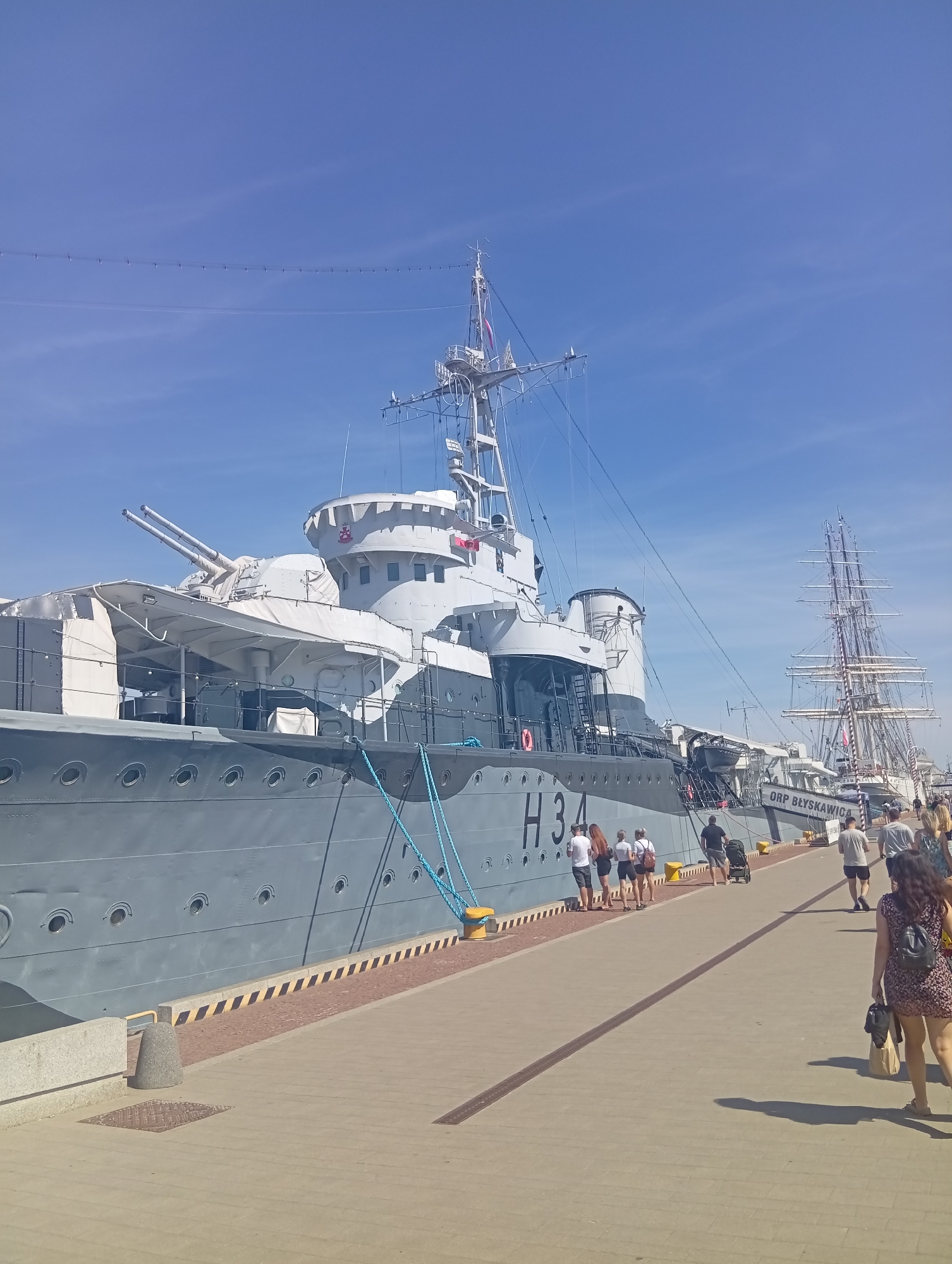 The image shows a daytime view of the ORP Błyskawica, a preserved Polish destroyer, docked in Gdynia, Poland. The ship is painted in a grey camouflage pattern and has "H34" visible on its hull, along with "ORP BŁYSKAWICA" written on its side. Several people are walking along the pier in front of the ship, some are looking at the ship and some are walking away from the ship. In the background, a large sailing ship with multiple masts is also visible. The sky is clear and blue. The pier is made of concrete pavers with a patterned edge.