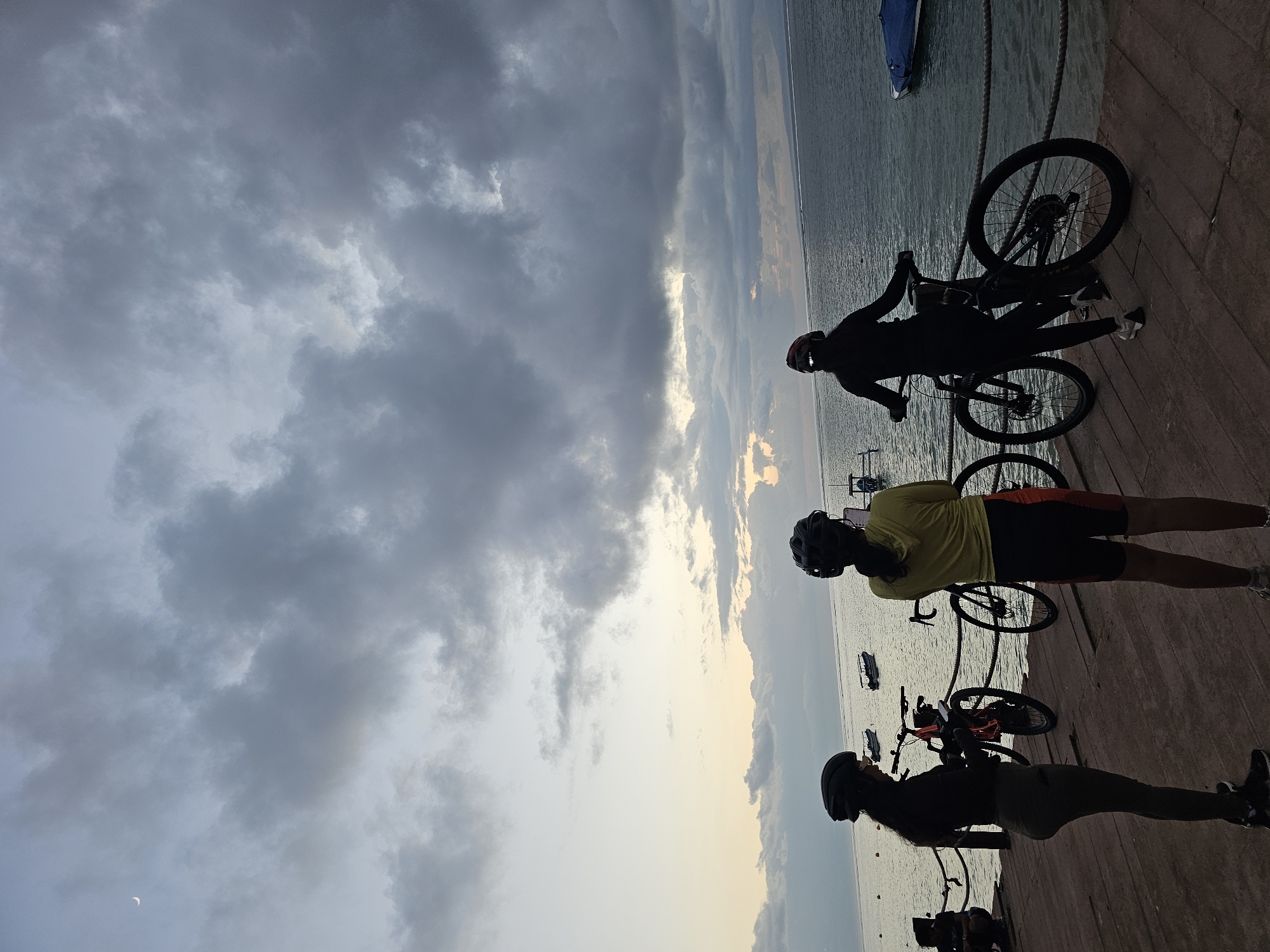 Three individuals wearing cycling helmets stand on a wooden pier next to their bicycles. The person on the left wears dark clothing and is looking down at their phone. The person in the middle wears a yellow hoodie and black shorts with orange accents and is looking towards the water. The person on the right wears black athletic clothing and is facing away from the camera, looking out at the sea. The sea stretches out before them, with several small boats visible in the distance. The sky above is filled with dramatic, dark clouds, with a sliver of the crescent moon visible in the upper left corner. The scene appears to be set during either sunrise or sunset. The wooden pier is weathered, and a rope railing runs along the edge. The location is likely a coastal area in Denpasar, Indonesia, given the presence of boats and the tropical atmosphere suggested by the lighting and vegetation visible in the distance.