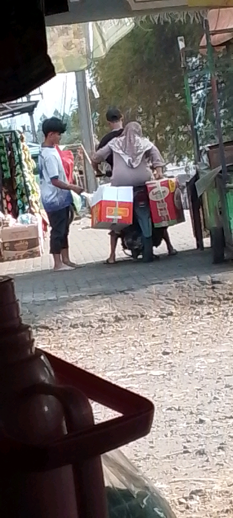 In the city of Cilaku, Indonesia, a young person stands on a paved walkway, holding a red object. They are wearing a white t-shirt with blue markings and dark pants. Behind them, two individuals are seated on a motorcycle. The person in front wears a black cap and a dark shirt, while the person behind them wears a light-colored hijab and a light brown garment. The motorcycle carries two large boxes, one white with orange trim and the other red with white lettering. A stall with merchandise is visible to the left, and greenery and trees are present in the background. The ground is covered with gravel and loose stones. The scene appears to be outdoors, possibly a market or roadside stall.