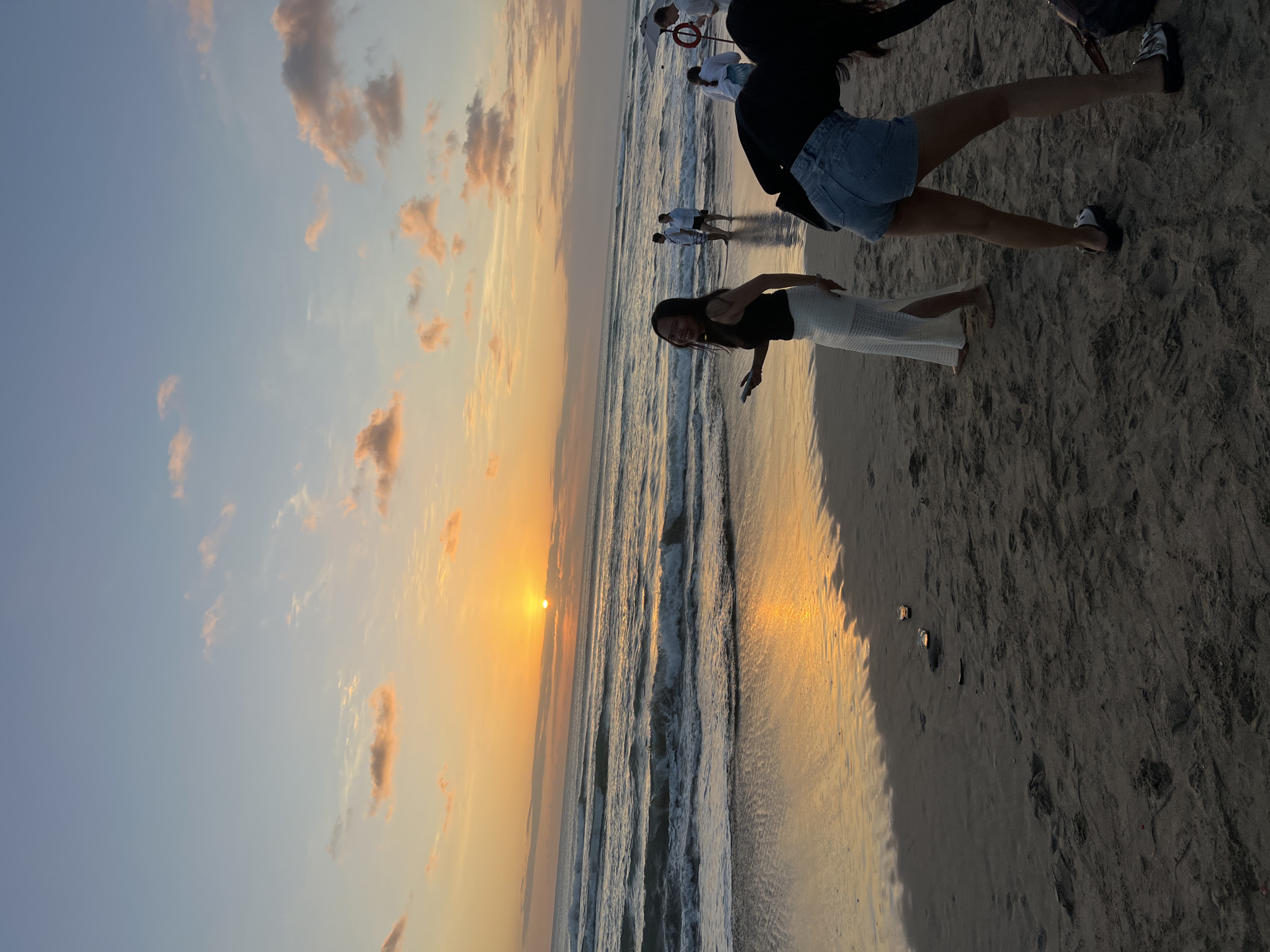 The image captures a sunset scene on the beach in Kuta, Indonesia. The sun is setting over the ocean, casting a warm orange glow across the sky and reflecting on the wet sand. Waves are breaking on the shore, creating a tranquil atmosphere. Several people are present on the beach, including two women in the foreground. One woman, wearing a black top and a white skirt, is smiling and holding a phone. The other woman, in a black top and denim shorts, is bending over. In the background, other beachgoers can be seen enjoying the sunset. The sand is a dark, wet color, with footprints visible. There are a few small objects on the sand near the water's edge, possibly discarded items. The overall scene is one of relaxation and natural beauty.