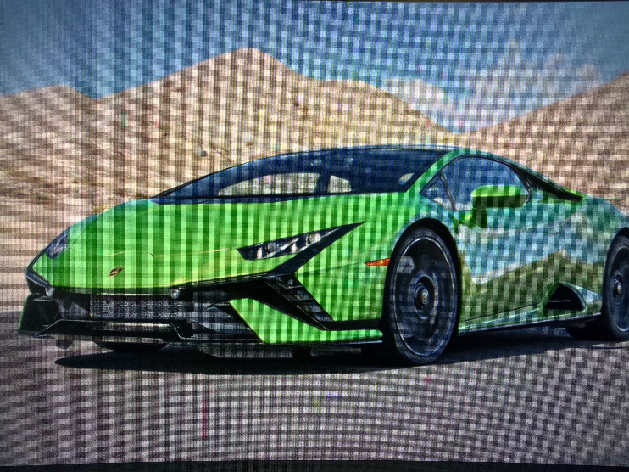 The image displays a vibrant lime green Lamborghini Huracán EVO supercar, positioned prominently on an asphalt road. The car, with its sleek, aerodynamic design and black accents on the front bumper and side skirts, appears to be in motion, as suggested by the slight blur on its wheels. A small Lamborghini emblem, featuring a charging bull, is visible on the front hood.
The setting is an outdoor, arid environment under a bright, clear blue sky with sparse white clouds. In the background, large, sandy-colored mountains or hills dominate the horizon, creating a vast desert or mountainous landscape. The lighting indicates it is daytime, likely late morning or early afternoon, with sharp shadows cast beneath the car. There are no people visible in the image. No discernible text, such as a license plate, is clearly visible due to the pixelated nature of the image, which appears to be a photograph of a digital screen. The scene conveys a sense of speed and luxury within a striking natural backdrop.
