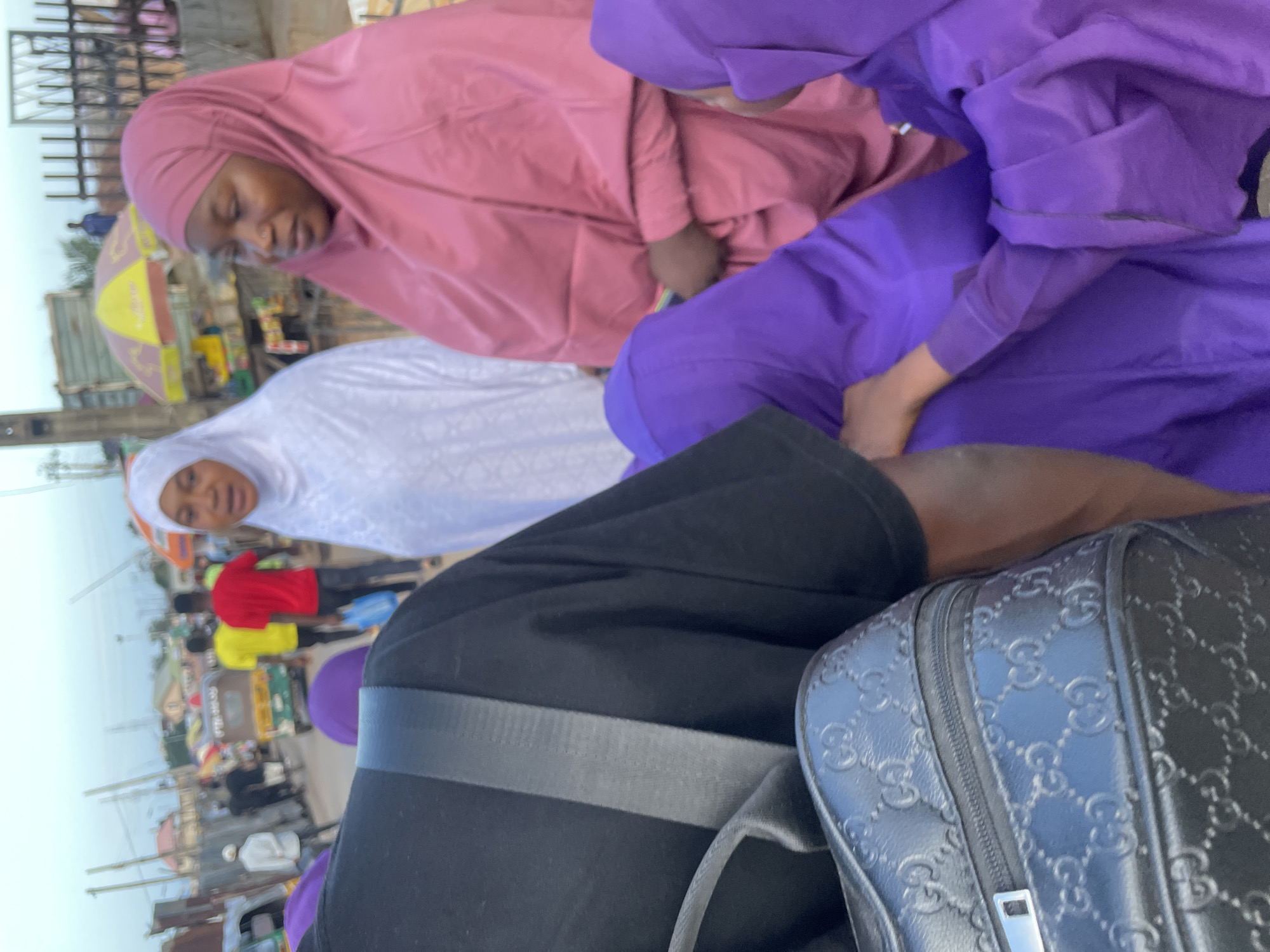 In Jos, Nigeria, a group of people are seen on a street, likely a market. In the foreground, a person wearing a black shirt and carrying a black Gucci-embossed bag is partially visible, facing away from the camera. Two women in hijabs are prominent subjects; one wears a pink hijab and the other a white hijab. Behind them, two men in brightly colored shirts, red and yellow, are walking away from the camera, and a man in a white shirt is seen to the left. A yellow and red umbrella and various stalls are visible in the background, suggesting a bustling marketplace environment. The overall scene depicts everyday life in an urban Nigerian setting.