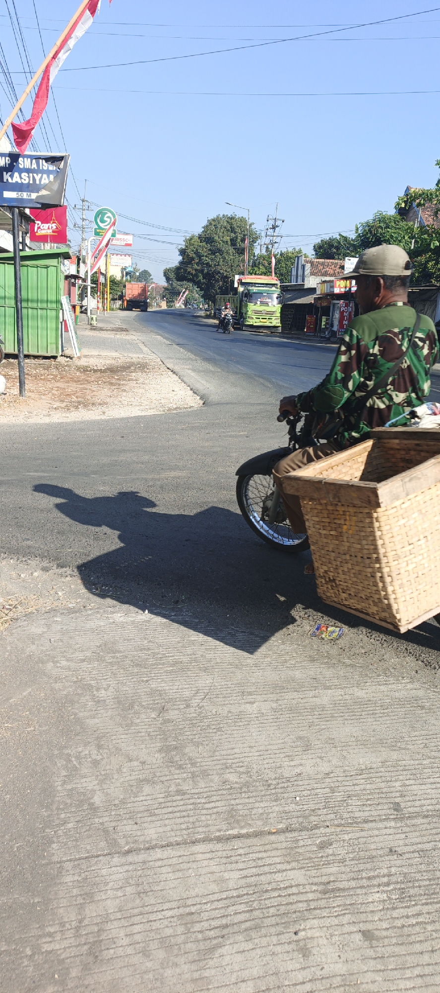 A man wearing a camouflage shirt and a cap rides a motorcycle with a large woven basket attached to the rear in Kasiyan, Indonesia. The man is on a paved road next to a sidewalk made of concrete with a grooved texture. The road has a shadow cast by the motorcycle and the rider. In the background, there are businesses with signs, including one that reads "MP SMA ISLAM KASIYAN" and another that says "Paris". There are also trucks and other vehicles on the road. Trees and buildings line the side of the road. The sky is clear and blue, indicating a sunny day.