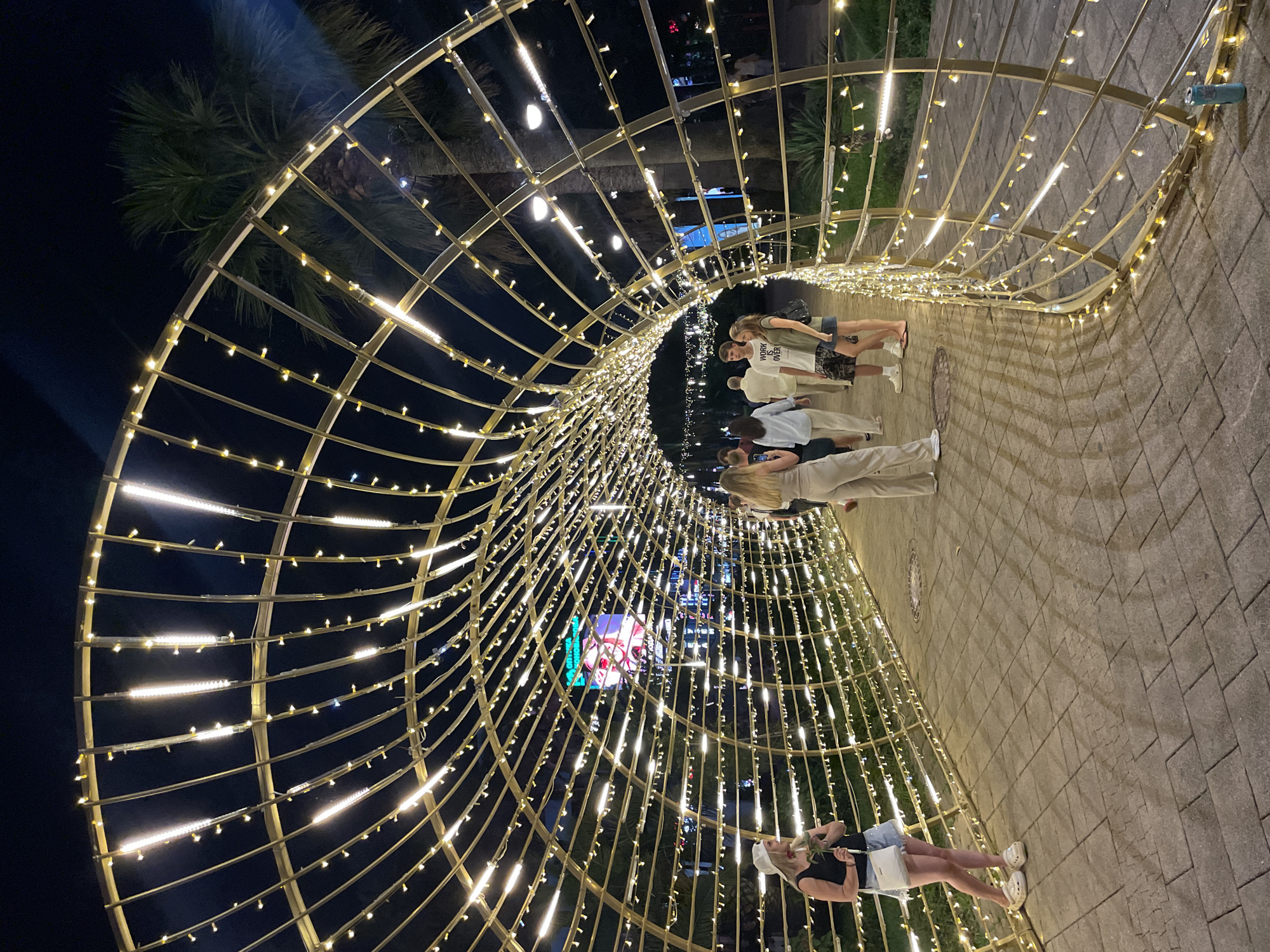 An outdoor nighttime scene in Sochi, Russia, features a tunnel-like structure adorned with numerous small, yellow lights and larger, white LED strips. The tunnel is constructed from a golden-colored metal frame with a curved, arched design. The ground within the tunnel is paved with interlocking stone blocks, and shadows are cast by the lighting structure.
Several people are visible walking through and around the illuminated tunnel. On the left side, a woman wearing a black tank top, denim shorts, and white Crocs stands holding a red object, possibly a rose. Further into the tunnel, a group of individuals, including a man in a white t-shirt with text and a woman in light-colored pants, are walking. On the right side of the tunnel's exit, two people, a man and a woman, are walking away from the camera. In the background, a palm tree is visible, and a large, brightly lit billboard displaying an image is present. The overall atmosphere suggests a vibrant, illuminated evening setting.