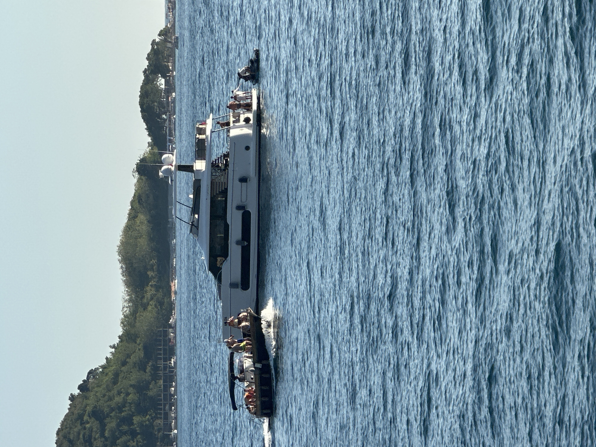 The image depicts a panoramic view of the sea off the coast of Budva, Montenegro, on a clear, sunny day. In the foreground, the bright blue water exhibits gentle waves. Dominating the middle ground are two yachts. The larger yacht, white and modern, is positioned to the right, with several individuals visible on its deck and near its stern. To its left, a smaller, darker boat, also carrying passengers, moves through the water, creating a wake. In the background, a lush, green hillside slopes down towards the sea, dotted with trees and some structures, suggesting a coastal town or resort area. The sky is a pale, cloudless blue.