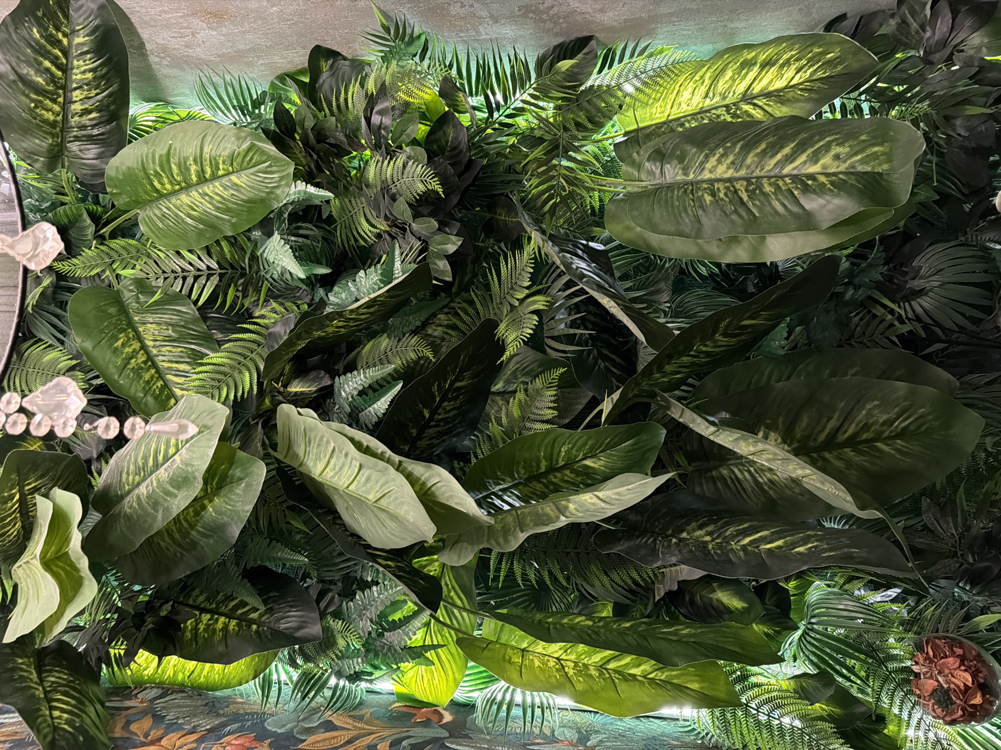 The image depicts a dense artificial plant arrangement, likely used for decorative purposes. Various types of foliage are visible, including large, broad leaves with mottled green and yellow patterns, as well as ferns and other leafy plants. The overall color scheme is dominated by shades of green, with some areas illuminated by artificial lighting. In the lower left corner, a decorative element resembling a white floral arrangement or a string of lights is visible. In the lower portion, a surface patterned with floral and botanical motifs can be seen. The location is not determinable from the image itself.