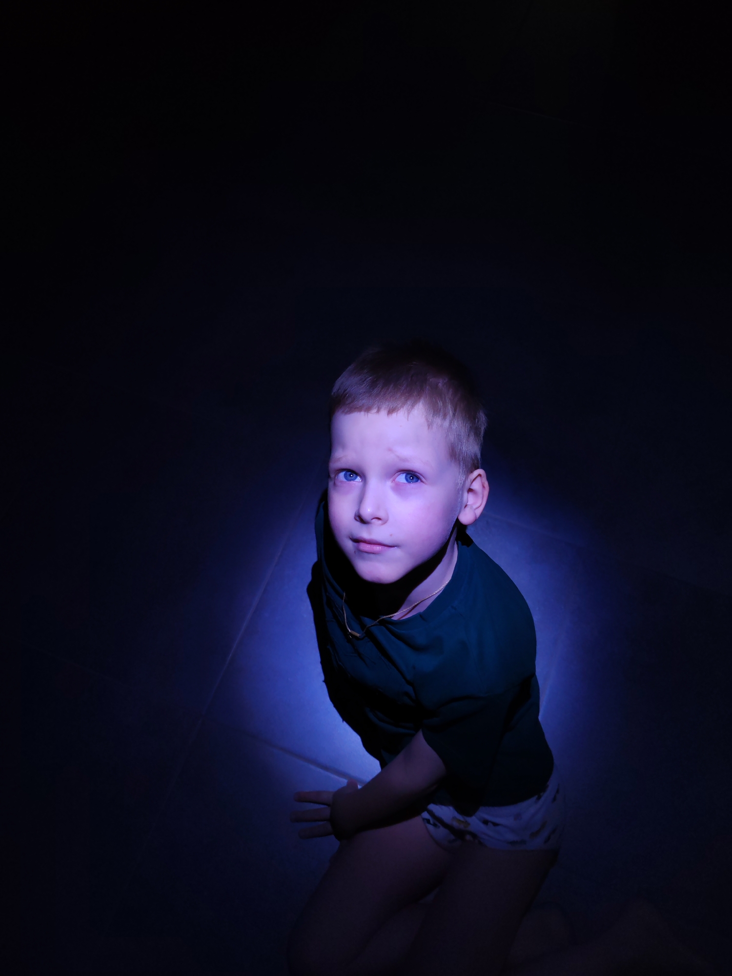 A young boy, likely between 5 and 8 years old, is the sole subject of the image, captured from a high-angle perspective. He is kneeling or sitting on the floor, looking upwards and slightly to his left with a direct, blue-eyed gaze.
The scene is dramatically lit by a single, strong, cool-toned (blue/purple) spotlight originating from above, illuminating his face, upper body, and a small, circular area of the floor beneath him. The rest of the environment is enveloped in deep shadow, creating a stark contrast and an intimate, focused atmosphere.
The boy is wearing a dark green short-sleeved t-shirt and light-colored shorts with a subtle, dark pattern. A thin, light-colored chain is visible around his neck. His expression is neutral, appearing somewhat contemplative or curious under the intense, colored light.
The setting is an interior space, likely a darkened room, possibly at night, given the precise and isolated nature of the illumination. No specific location cues are visible beyond the implied indoor environment. No text is present in the image. The location context for this media file is Chaikovsky, Russia.