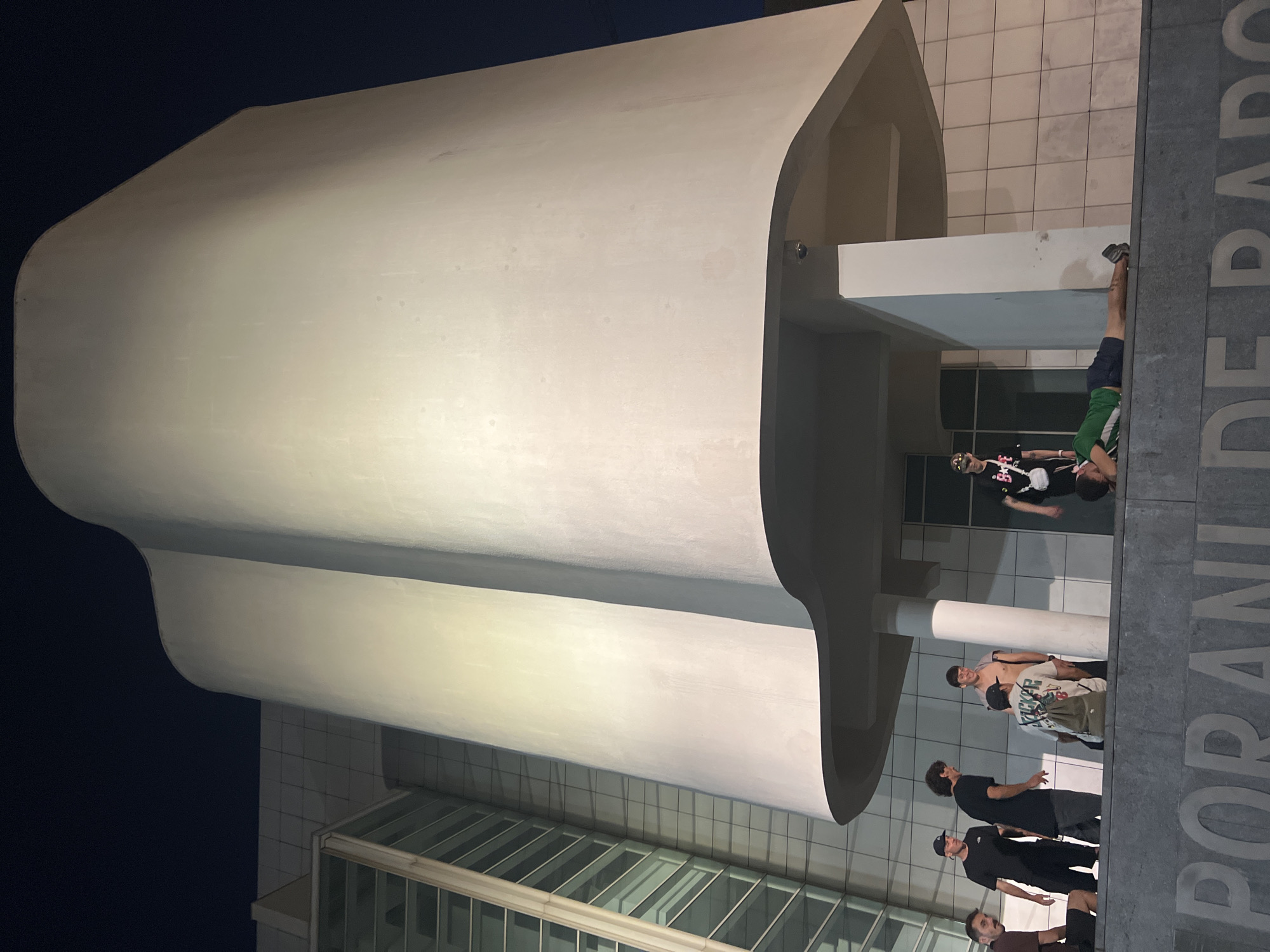 The image shows a group of young men gathered in front of a modern building at night. The building has a distinctive curved facade and is illuminated by artificial light. Several men are standing and talking, while one man is lying down on a ledge. The text "PORANIDE RADO" is visible on a stone wall in the foreground. This location is the MACBA (Museum of Contemporary Art of Barcelona) plaza in Barcelona, Spain, a popular gathering spot for young people and skaters. The atmosphere appears casual and relaxed, with the group interacting amongst themselves.