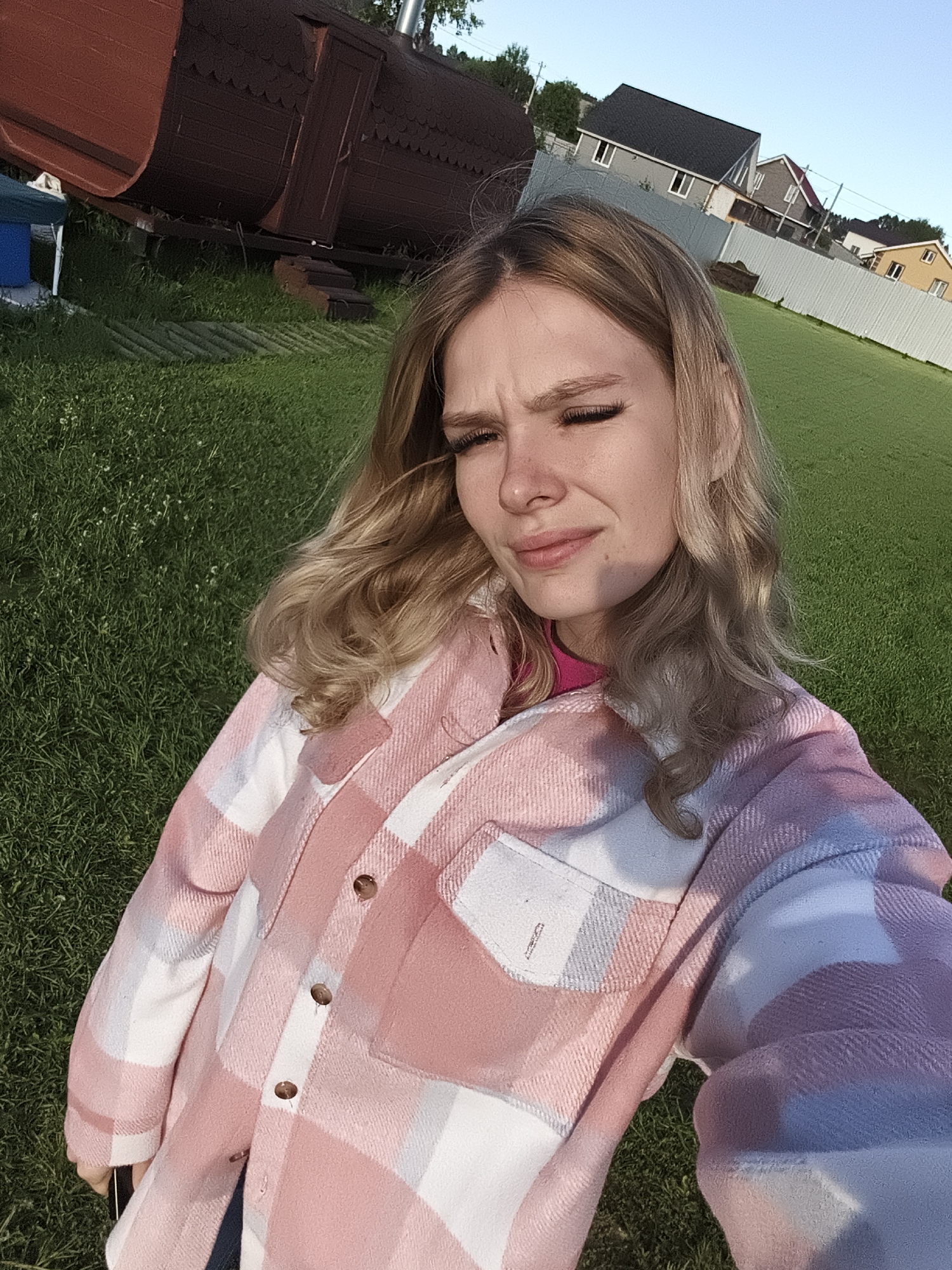 The image features a young woman taking a selfie outdoors in what appears to be a residential or rural setting in Русский Вожой, Russia. She has long, wavy blonde hair and fair skin. She is wearing a pink and white plaid, flannel-style jacket over a bright pink top. Her face is slightly turned, and she is squinting or grimacing, likely due to the bright, late afternoon or early evening sun, which casts a soft glow on her features. Her eyelashes are prominent.
In the background, to the left, is a distinctive dark reddish-brown barrel-shaped structure, possibly a sauna or a small hut, with a chimney visible on top. A partially visible blue object rests near its base. A light-colored corrugated fence stretches across the midground, separating the immediate grassy area from a row of houses. These houses exhibit varied architectural styles and colors, including grey, yellow, and white exteriors, with darker roofs. The ground is covered in lush green grass, suggesting a well-maintained yard or field. The sky above is clear and light blue, indicating good weather. There is no visible text in the image.