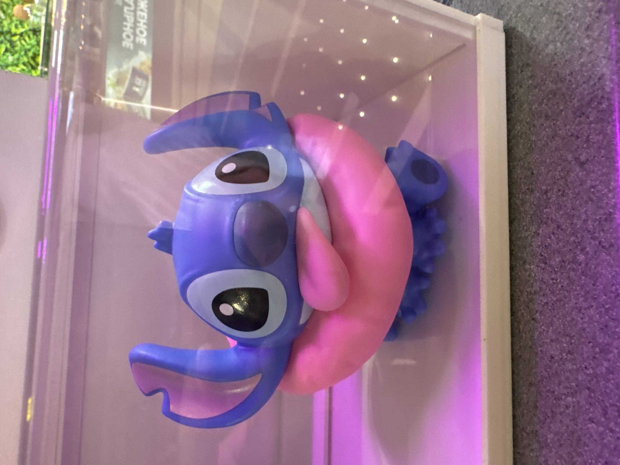 The image shows a blue and pink Stitch figurine displayed inside a white, illuminated display case. The figurine is primarily blue with pink accents. The display case has small lights inside, creating a slightly blurred background. The overall setting appears to be an indoor environment. The location is not discernible based on the provided image.