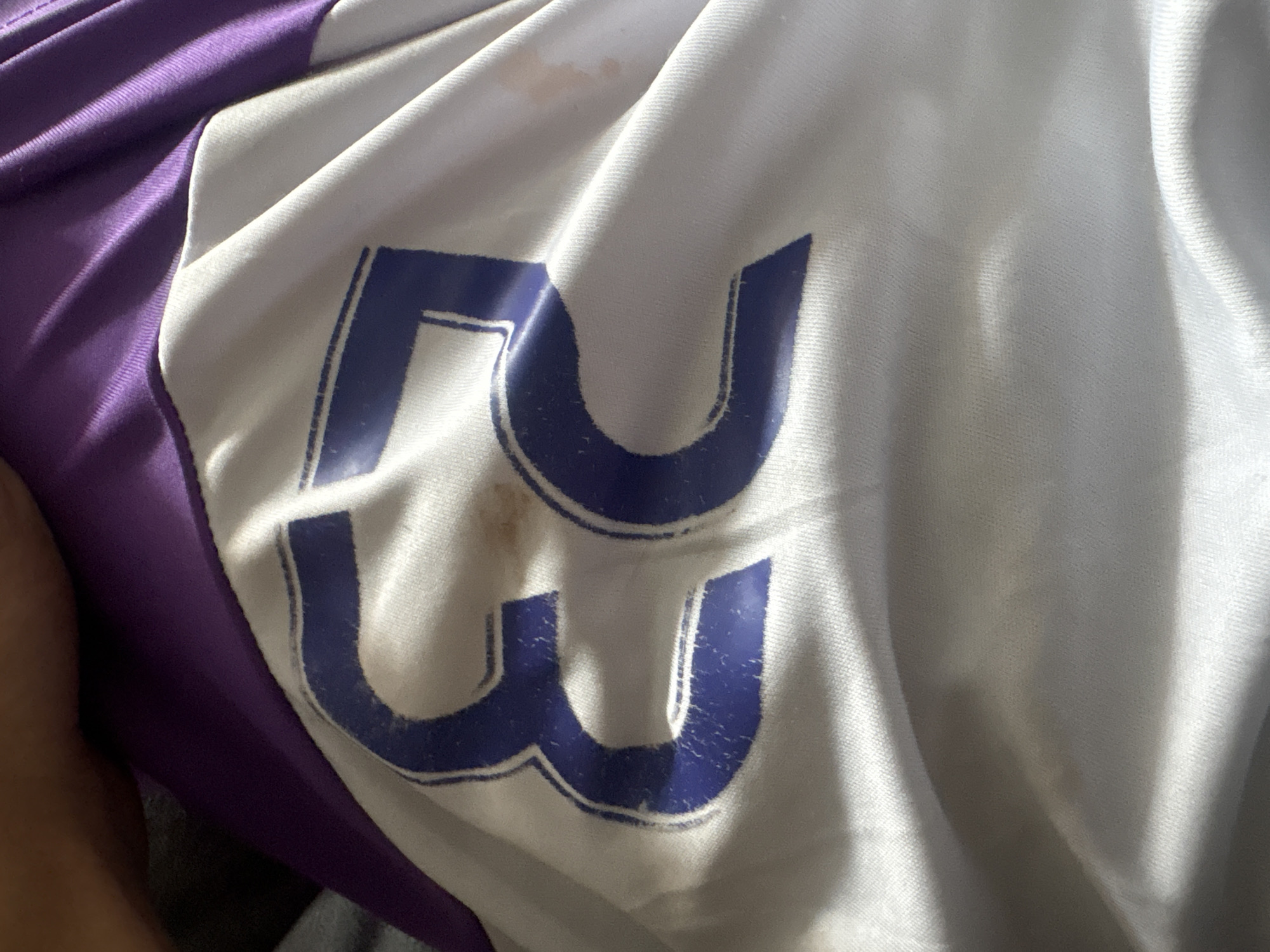 The image is a close-up, angled shot of a white fabric garment with purple trim along the top edge. A large blue number "53" is printed on the white fabric. There are several small brown and pinkish stains on the white fabric, as well as some wear and tear on the blue print of the number. The fabric is creased and folded, indicating it is likely a piece of clothing. There is no visible indication of the city of Petropavl, Kazakhstan in the image.