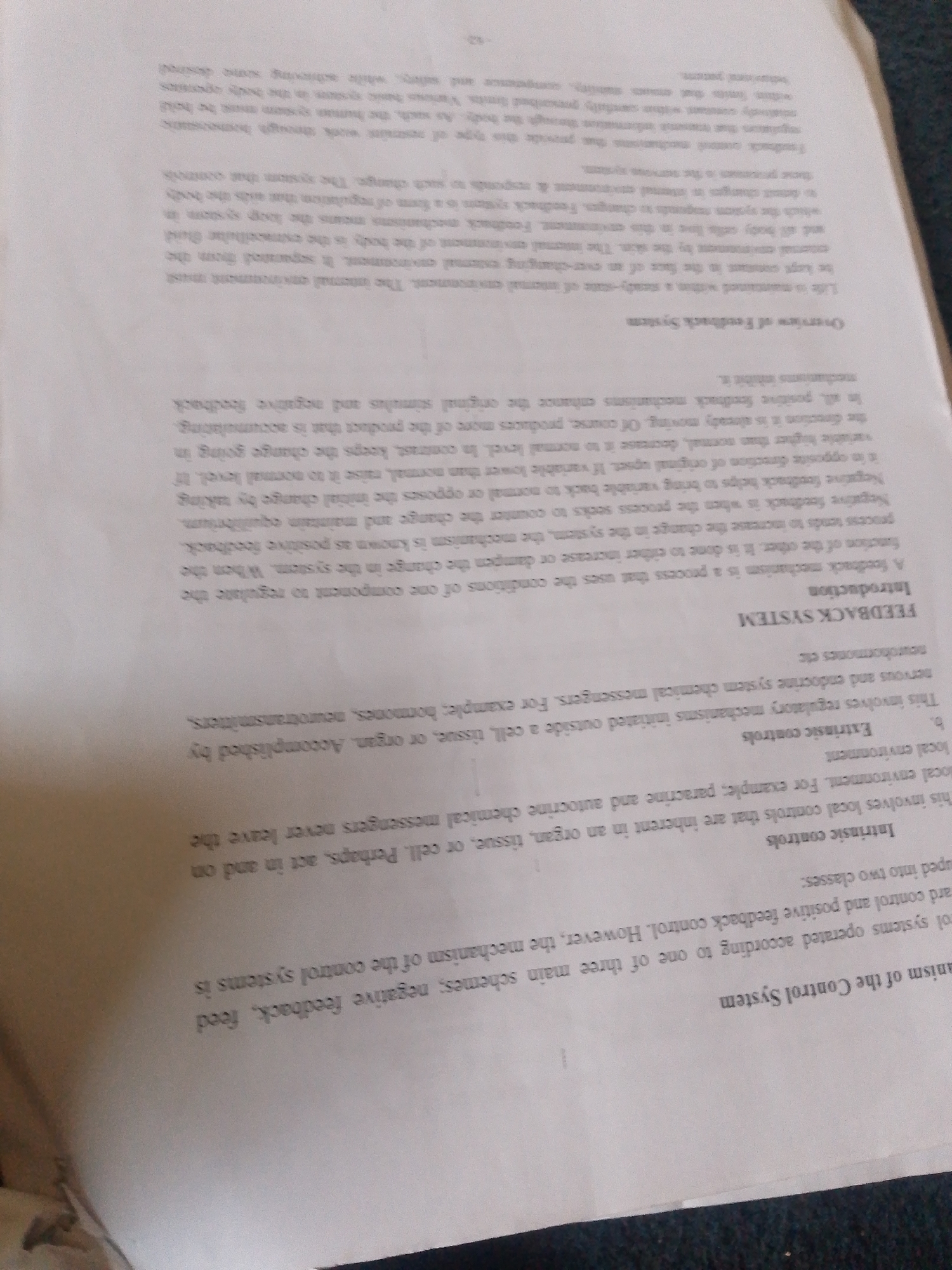 The image displays a close-up, angled shot of a book or document with text. The primary subject is the printed words on the pages. The text is predominantly in English and discusses "Feedback Systems" and "Control Systems." Specific sections mentioned include "Introduction," "FEEDBACK SYSTEM," "Intrinsic controls," and "Extrinsic controls."
The lighting suggests an indoor environment, possibly under artificial light, as there are no strong shadows or indications of natural daylight. The page texture appears to be standard paper, and the printing is black ink. The angle of the shot creates a slightly distorted perspective, with some lines of text appearing curved.
There are no people, objects, or discernible scene elements beyond the book itself. The activity is passive, simply presenting information. No specific time of day, weather, or location cues like "Jalingo, Nigeria" are visible within the image content itself. There are no discernible emotions conveyed by the image. The visible text is the most notable detail, offering a glimpse into a scientific or technical subject.