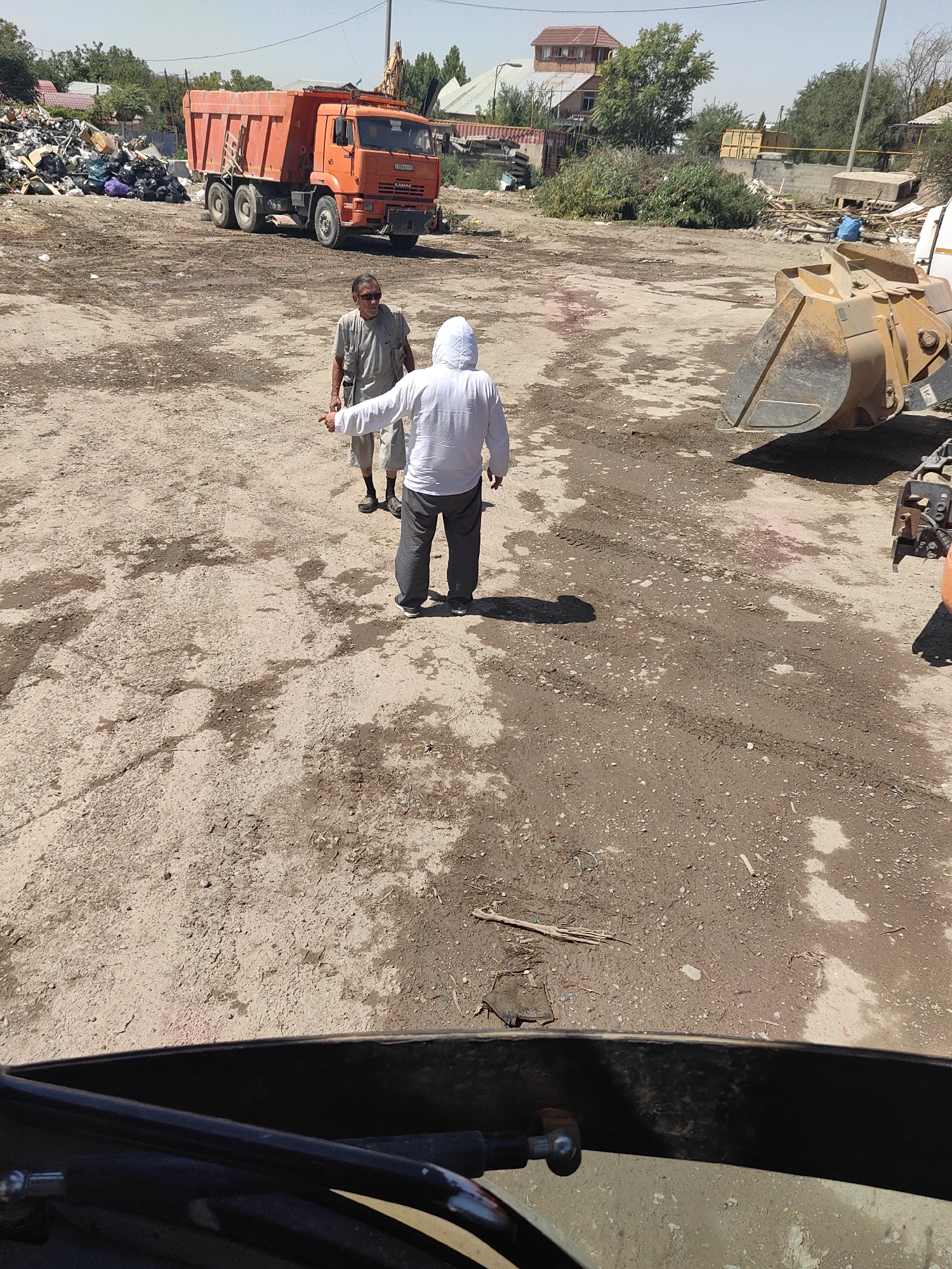 The image shows an outdoor scene in Shymkent, Kazakhstan, likely a junkyard or construction site. In the foreground, a person wearing a white hooded jacket is standing and pointing. To their left, another person in a gray t-shirt and shorts is also standing. Behind them, an orange dump truck is parked, and to the right, the bucket of a yellow excavator is visible. The ground is a mix of dirt, gravel, and puddles. In the background, there are piles of debris, some buildings, and trees. The sky is clear and the sun is shining.