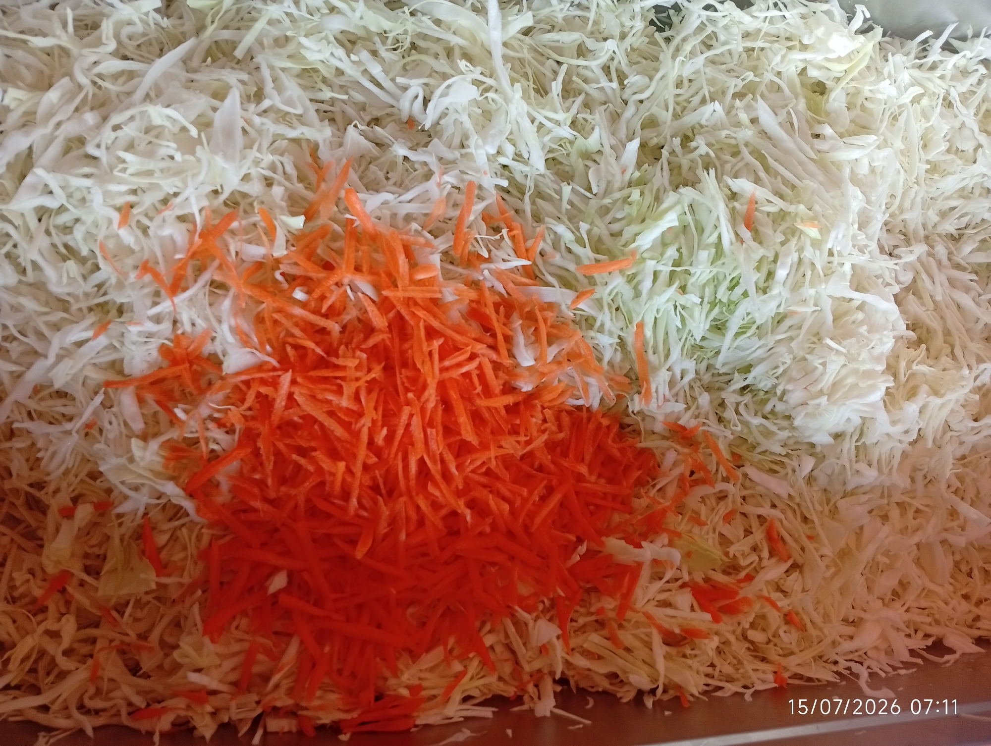 The image displays a close-up, top-down view of finely shredded cabbage, with a pile of shredded carrots concentrated in the center left of the frame. The shredded cabbage is predominantly white, with some lighter green strands visible. The carrots are a bright orange color. The ingredients appear to be in a shallow, metallic container, likely for preparation or storage. There are no people or other objects visible in the frame. The lighting is even, suggesting an indoor environment. A timestamp in the lower right corner reads "15/07/2026 07:11". The scene is located in Bana Stream, Nigeria.