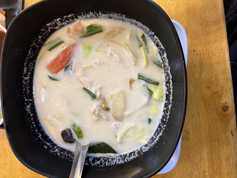 A black ceramic bowl, centrally positioned, contains a creamy white soup consistent with Thai Tom Kha Gai, indicative of Thai cuisine often found in Bangkok, Thailand. The specific establishment within the city is not discernible.
The soup is filled with various ingredients: sliced chicken pieces, dark brown mushrooms, bright red tomato wedges, pale green cabbage or leafy greens, elongated green spring onion or herb stalks, and light beige slices likely identifying as galangal or similar root. A silver-colored metal spoon rests on the left side of the bowl, partially submerged in the soup. The liquid exhibits some frothing along the inner rim of the bowl.
The bowl itself has a slightly textured or speckled appearance on its inner rim. It rests on a light-colored wooden table surface, displaying prominent grain and minor wear. A small white dish or coaster is partially visible beneath the bowl's lower left edge. The background, out of focus, shows hints of other dark tableware and reddish-orange elements. No individuals or specific actions are depicted.