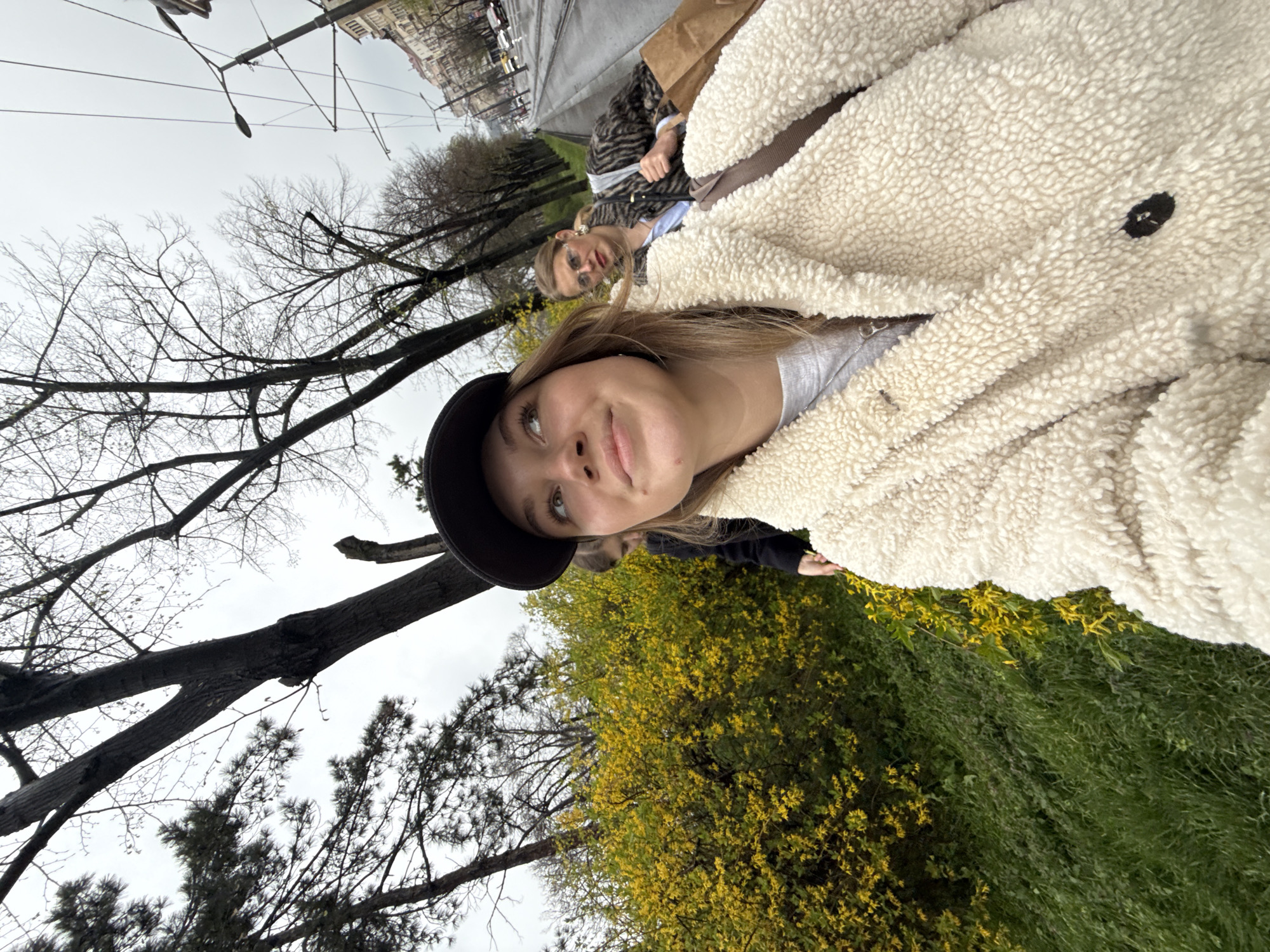 The image captures an outdoor selfie on a city street in Belgrade, Serbia, under an overcast sky during daytime. In the foreground, a young female subject is seen taking the photograph, looking upwards and to her right with a gentle smile. She wears a black baseball cap, a light grey t-shirt, and a cream-colored, textured "teddy" coat, with a silver chain necklace. A brown backpack strap is visible over her right shoulder. Behind her, slightly out of focus, a second female subject is visible, wearing sunglasses, a patterned coat (possibly animal print), and a light blue collared shirt. She carries a light brown paper bag in her left hand and a dark shoulder bag over her right. A third individual is partially obscured behind the primary subject. To the left, a dense bush with numerous bright yellow flowers (consistent with Forsythia) and green grass is prominent. The background features a wide city street or boulevard equipped with tram tracks and overhead electrical lines. Tall, bare deciduous trees line the street, interspersed with some coniferous trees on the far left. In the distance, multiple-story buildings and light vehicular traffic are visible along the boulevard.