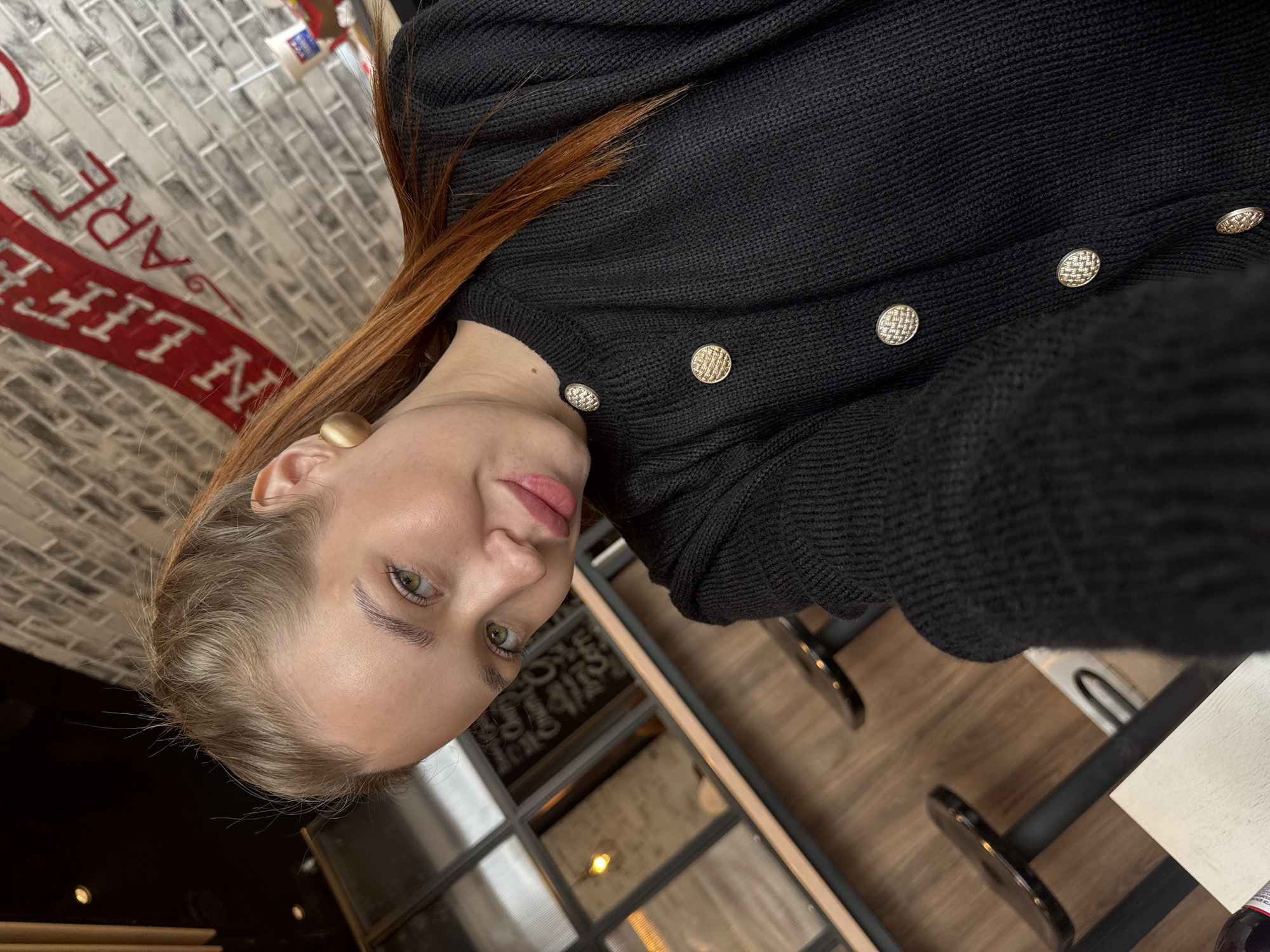 A young Caucasian woman with blonde hair pulled back in a ponytail and green eyes is taking a selfie. She is wearing a black knitted cardigan with gold buttons and large gold circular earrings. She is in a cafe with a brick wall in the background displaying a red banner with white lettering that reads "LIFE ARE". There are also several stools and a wooden counter visible. The overall lighting suggests an indoor setting, possibly during the day. The woman is looking directly at the camera with a slight smile.
