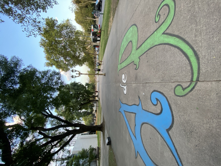 The image depicts a wide, concrete pedestrian pathway in the Belgrano neighborhood of Buenos Aires, Argentina, likely along Avenida Figueroa Alcorta in the vicinity of the Estadio Mâs Monumental.
In the foreground, large, stylized letters are painted directly onto the concrete path: a blue 'A' on the left, a small white 'de' in the center, and a green 'R' on the right. These letters face the viewer, oriented to be read looking forward along the path. The pathway extends into the midground, flanked by parallel lines of large, mature trees with dense green foliage. A multi-lamp, ornate black lamppost stands on the right side of the path.
In the midground, several individuals are walking along the path. One person walks alone on the left, further down. A group of three people walks together on the right side of the path, near the lamppost. Two additional figures are visible further down the path on the right. To the far left, a large, white building with visible structural elements, consistent with a stadium or public facility, is partially visible. A dark park bench is situated near this building.
The background reveals a paved street running parallel to the pedestrian path on the right. A red and black city bus is visible on the street, partially obscured by trees, along with a dark-colored car further back. The sky above is clear and bright blue with scattered white clouds, indicating daylight conditions. Sunlight appears to originate from the left, casting shadows from the trees and illuminating the right side of the path.