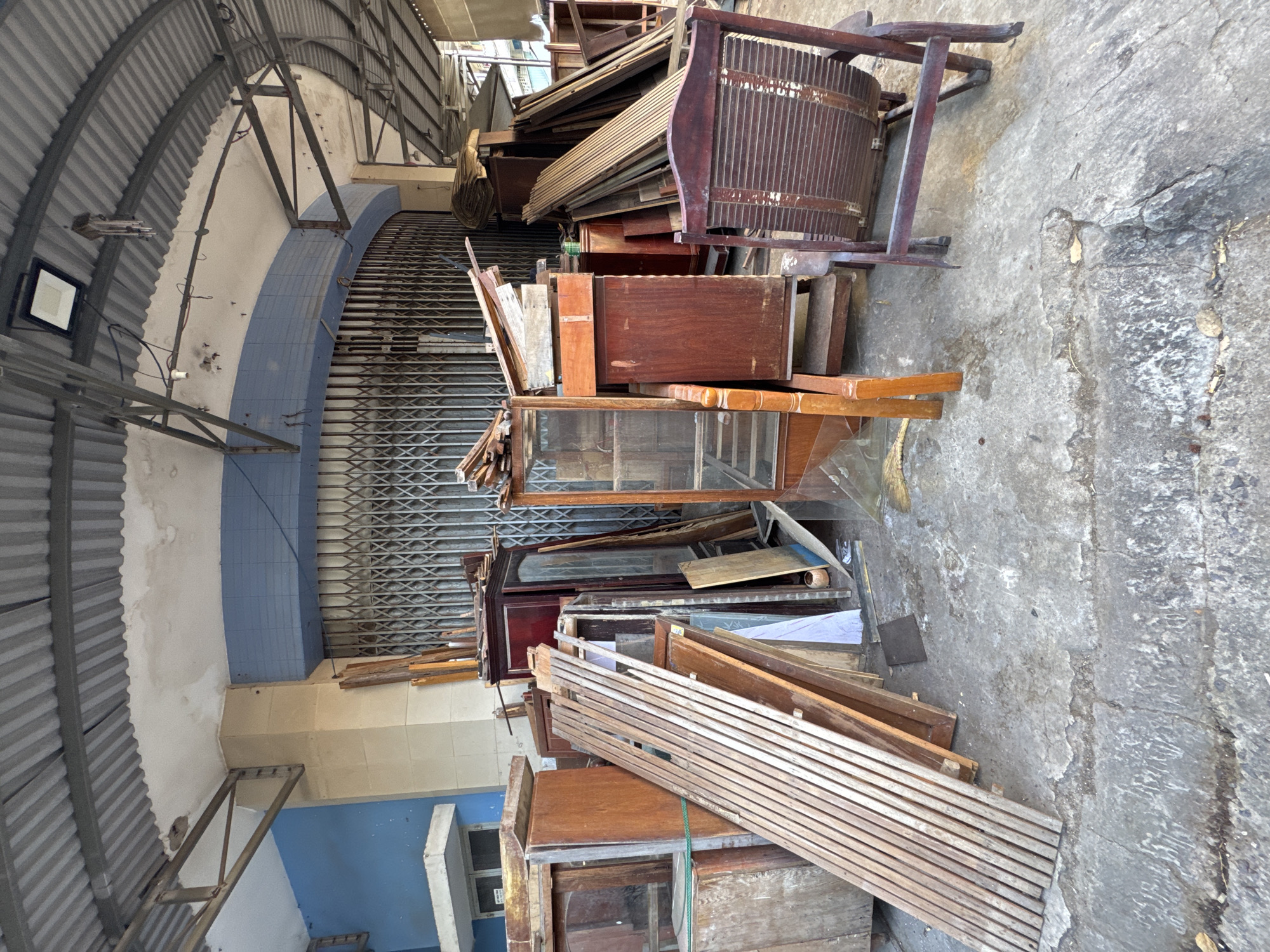 This is a wide shot of an outdoor area, likely in front of a shop or storage space, with various furniture items and building materials piled up. The foreground is a cracked concrete surface with debris and scattered leaves. Dominating the scene are wooden furniture pieces, including several tall cabinets, some with glass doors, and a wooden chair with a slatted back. Stacked wooden planks and other assorted wooden materials are leaning against the cabinets and piled on the ground. Behind the furniture, a large, rolled-up metal security gate is visible. Above, a corrugated metal roof is supported by exposed metal beams, creating an overhang. The background shows a blue tiled wall. The scene appears to be a storage or holding area for used or discarded furniture and wood. The location is specified as Nha Trang Ward, Vietnam.