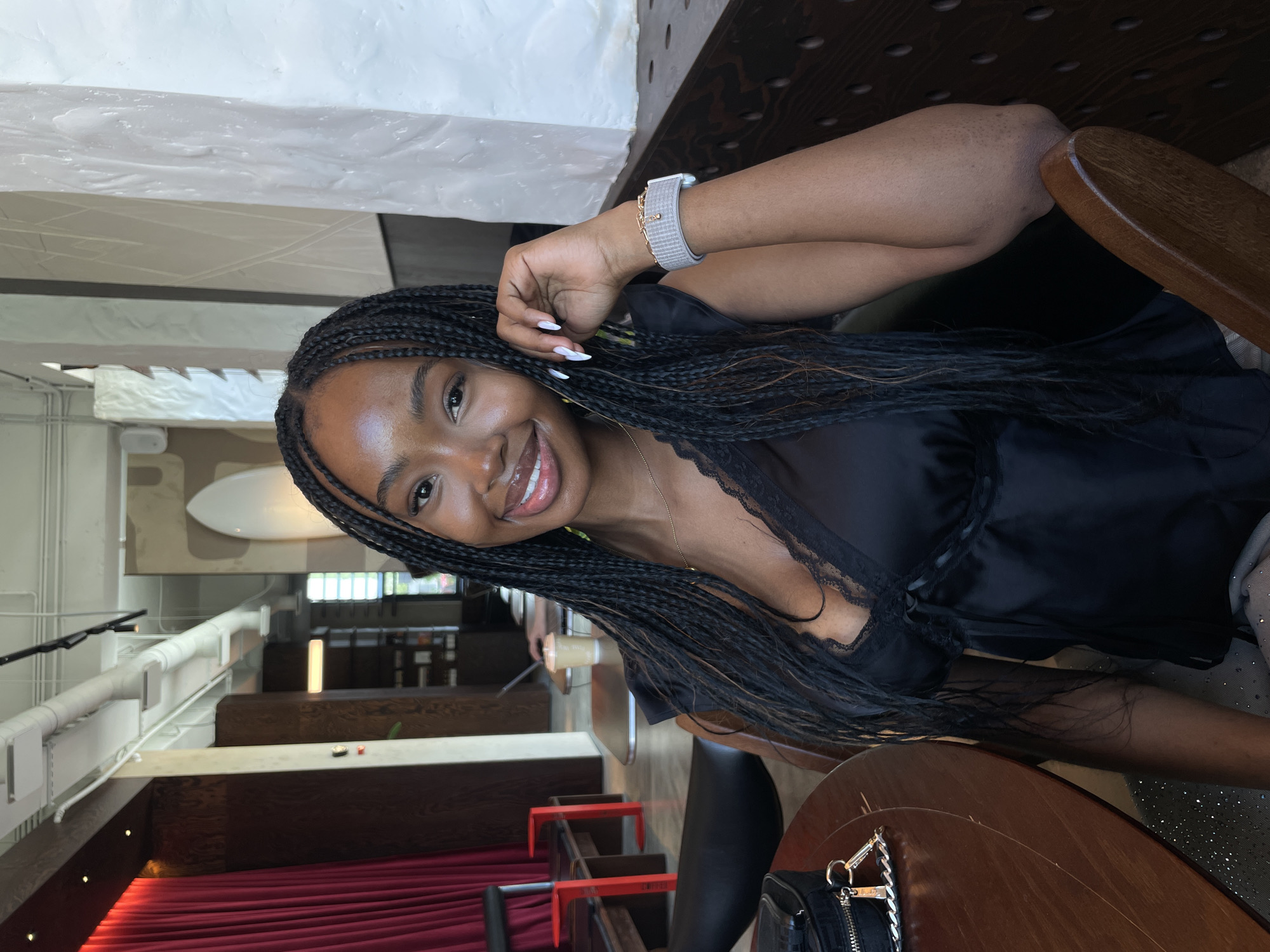 A young Black woman with braided hair is seated at a table in what appears to be a modern cafe or restaurant. She is smiling and looking towards the camera. She is wearing a black top with lace trim and a silver smartwatch. In the background, there are other tables, chairs, and decor, including red curtains and abstract art. The environment suggests an indoor seating area with ambient lighting. The city of Kazan, Russia, is not discernible from the image.