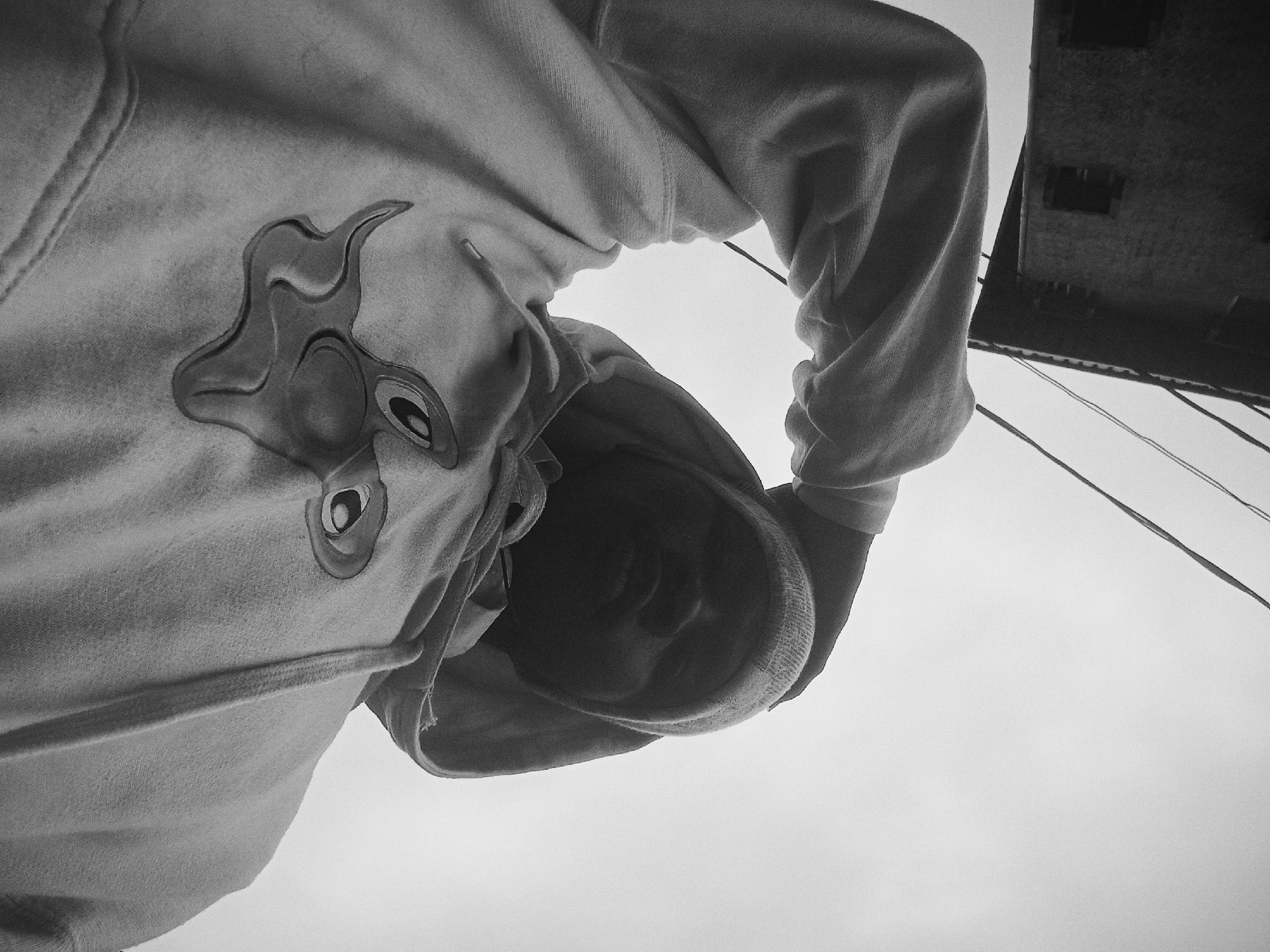 A low-angle, grayscale photograph shows a person wearing a hooded sweatshirt. The person is positioned above the camera, looking down. A cartoon-like design is visible on the front of the sweatshirt. In the background, partially visible in the upper right corner, is a building with wires running in front of it, and the sky. The city of Nkpor, Nigeria is not discernable from the image.