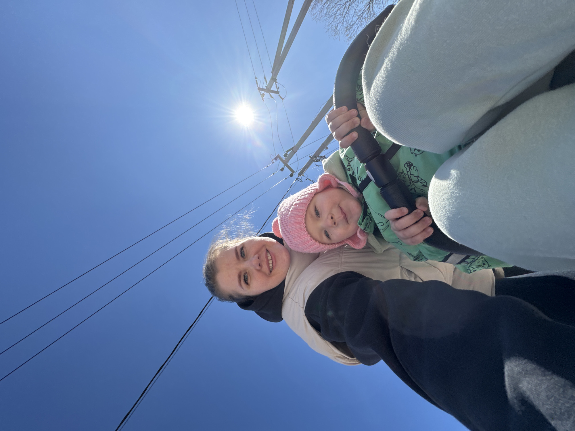 A low-angle shot shows a woman and a baby outside on a sunny day. The woman has fair skin, light hair pulled back, and is smiling at the camera. She wears a black hooded sweatshirt and a beige vest. The baby is next to her in what appears to be a stroller or baby carrier. The baby wears a light pink hat and a light green jacket. Power lines are visible against the clear blue sky, with the sun shining brightly in the upper left portion of the image. The location is identified as Горный, Russia.