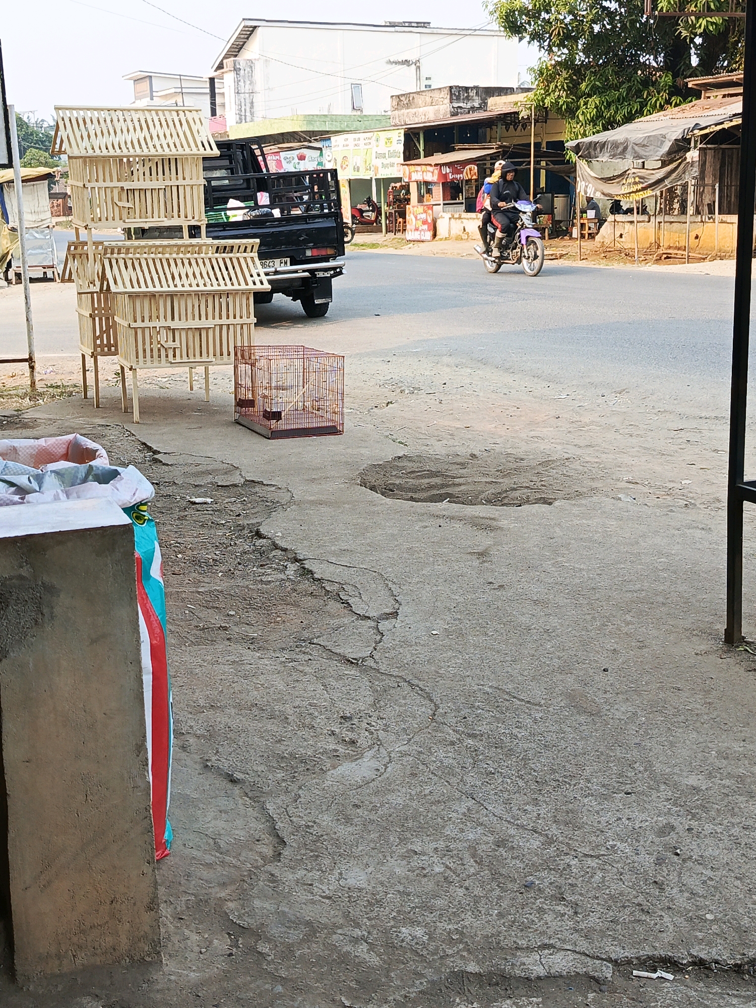 The image depicts a street scene in Megang Sakti I, Indonesia. In the foreground, to the left, is a concrete structure with a large bag partially visible, featuring a red, white, and teal striped pattern. Adjacent to this, to the right, are several wooden bird cages stacked and displayed. One smaller, orange metal bird cage is also present on the ground in front of the wooden cages. Further back, a black pickup truck is parked. The street is paved, with visible cracks and patches of discolored concrete, indicating some wear and tear. On the right side of the street, a row of storefronts with corrugated metal roofs is visible. A purple motorcycle with two occupants, a driver and a passenger, is traveling on the road, moving away from the viewer. The passenger is wearing a dark hooded jacket and the driver is wearing a dark jacket and pants. The background shows buildings, trees, and utility poles under a clear sky. The overall scene suggests a commercial area with outdoor displays of goods.