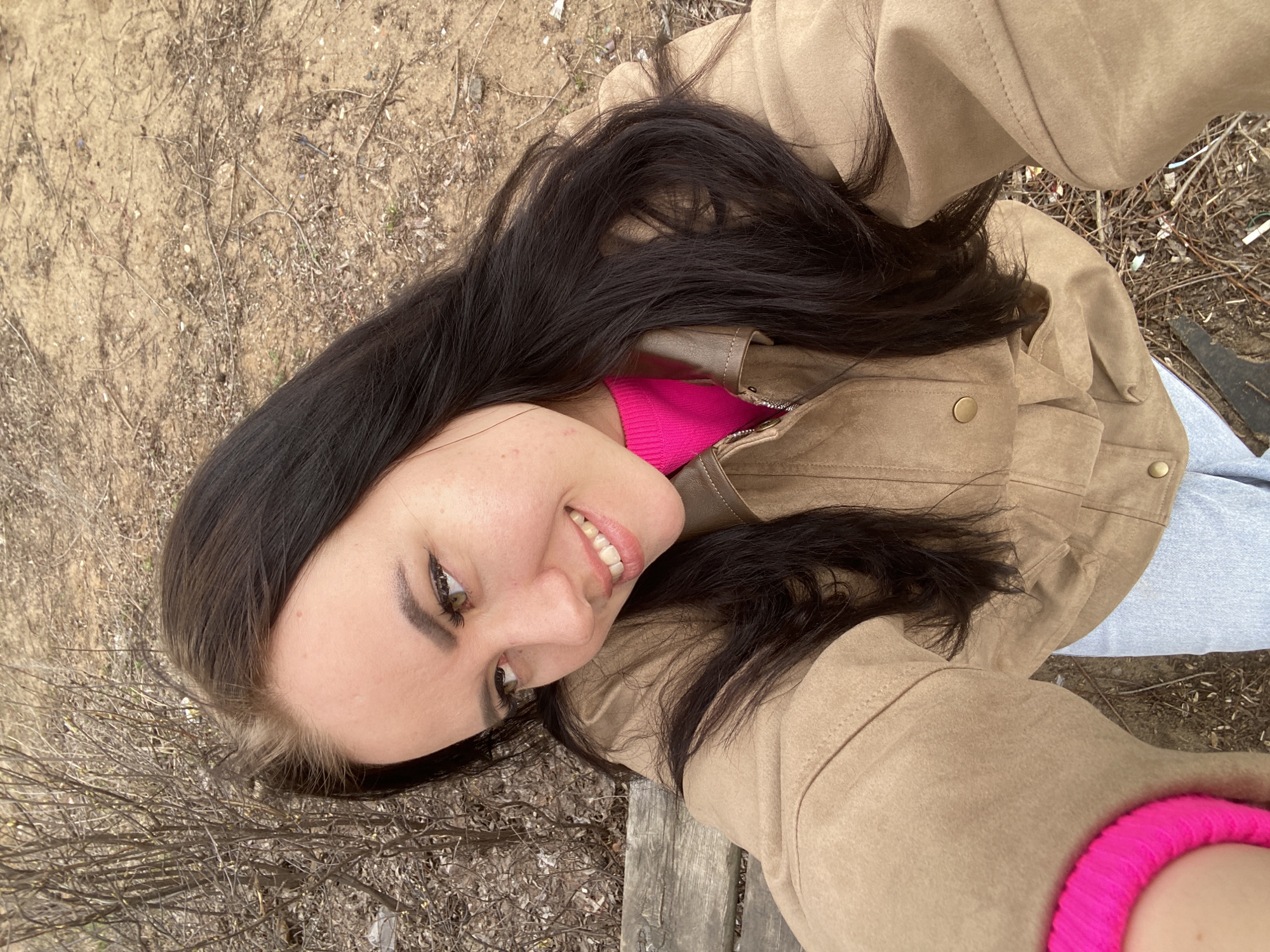 A selfie photograph taken outdoors, possibly in or near Chaikovsky, Russia, shows a young woman with dark hair and fair skin. She is wearing a bright pink top under a light brown jacket. The background is a mix of bare earth and vegetation, suggestive of a park or other natural area. The angle is from above, tilted, indicating the woman is holding the camera at arm's length. The lighting suggests it is daytime.