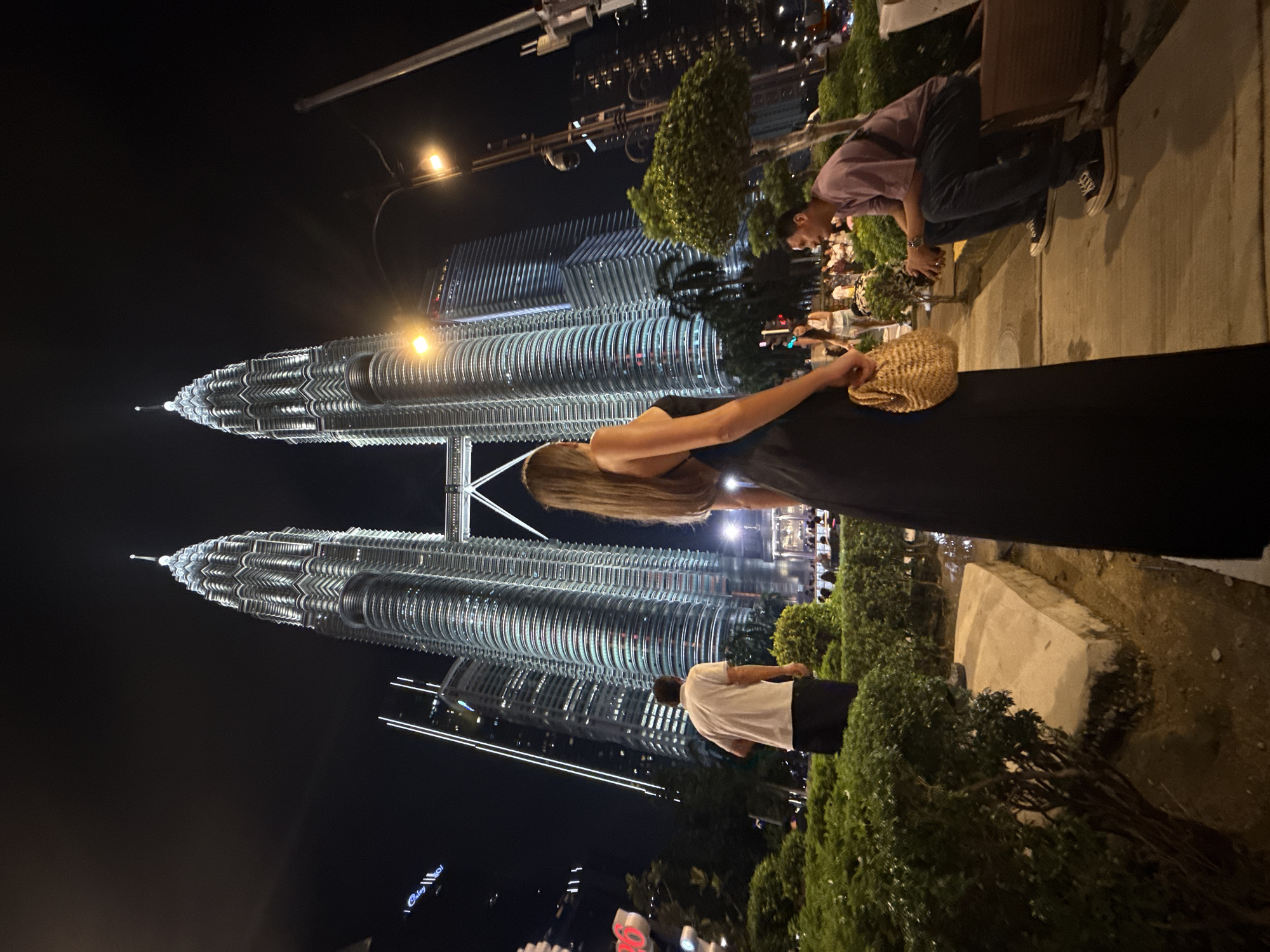 The image shows a night scene in Kuala Lumpur, Malaysia, featuring the Petronas Twin Towers illuminated against a dark sky. In the foreground, a woman in a black dress holds a straw bag. To the right, a man wearing a purple shirt and dark pants crouches near some greenery. Nearer to the middle-ground on the right, a man in a white shirt and black pants can be seen among the greenery. There are decorative lights and pedestrian traffic in the area. The environment is a public space, possibly a park or promenade, with trees and landscaped areas.