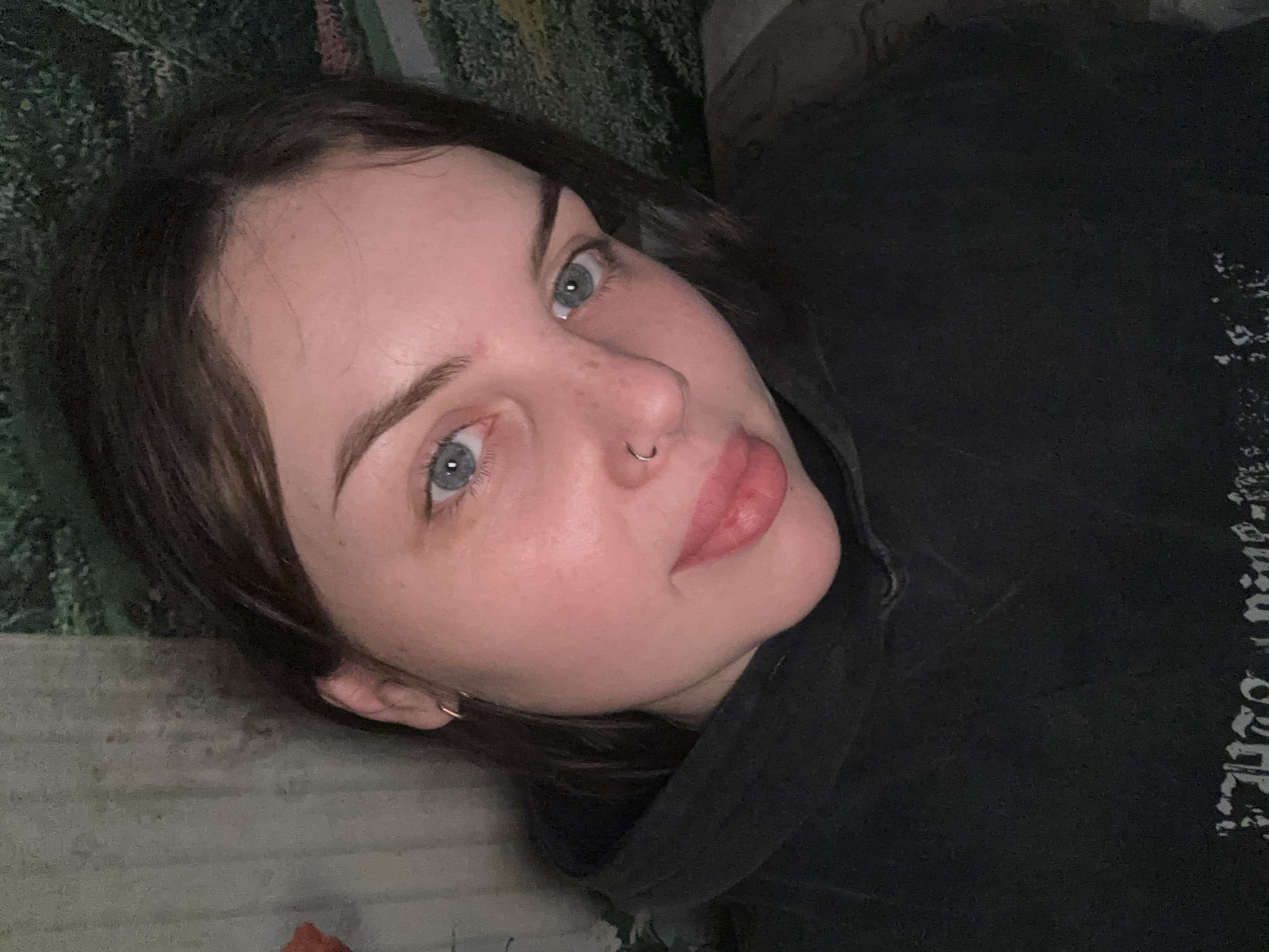 A close-up, low-angle shot shows a woman lying down, facing the camera with her head slightly tilted. She has fair skin, dark hair, light blue eyes, and a small nose ring. She is wearing a dark hooded sweatshirt. The background behind her head is blurred, showing patches of green and hints of an indeterminate pattern. The location is not identifiable as the city of Chornomorskoe, Ukraine, from the given image.
