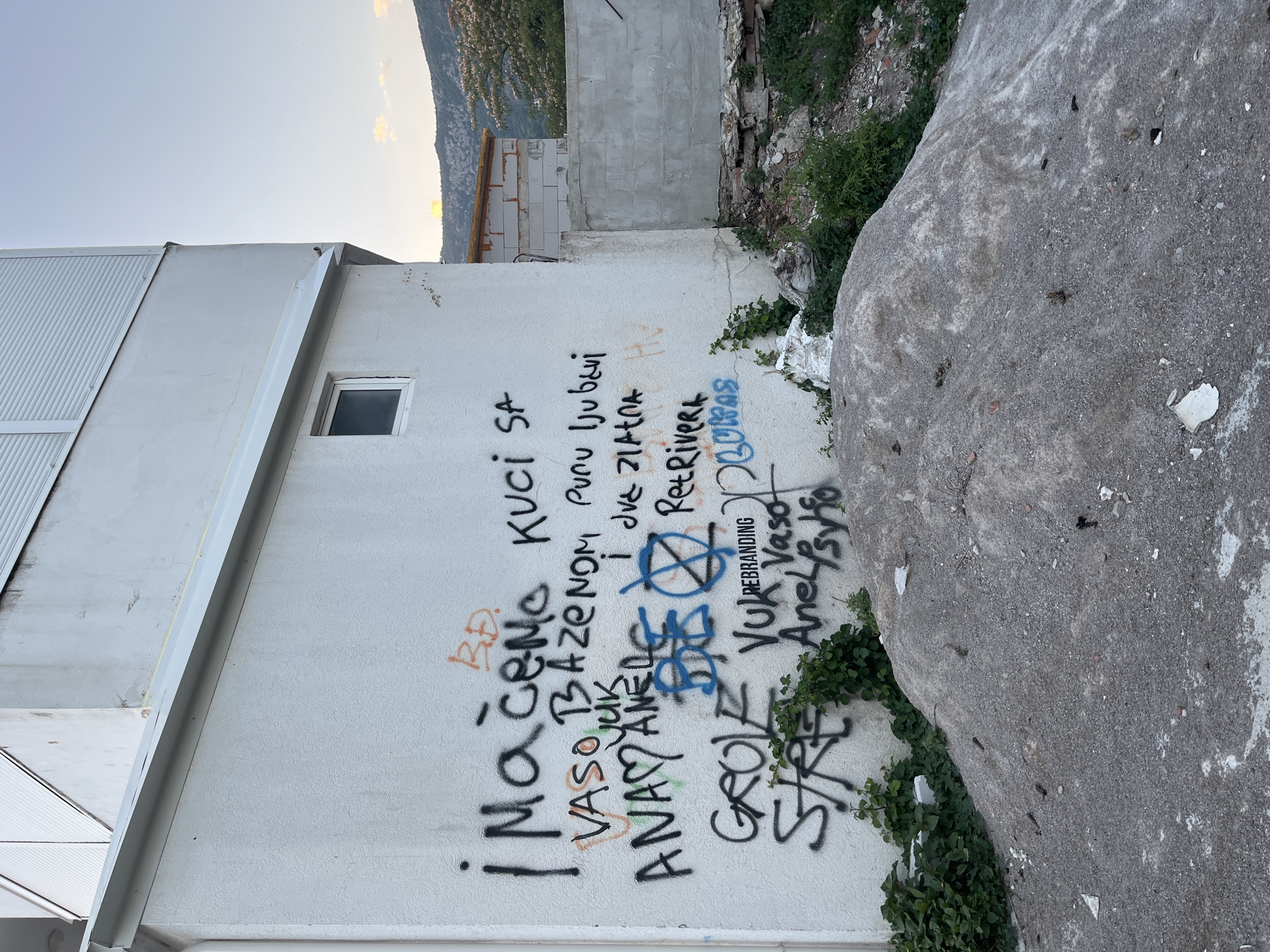 In the city of Budva, Montenegro, a white stucco building with a light gray roof overhang is covered in various graffiti. The graffiti includes black spray-painted words such as "¡Mačemo", "KUCI SA", "VASO BAZENOM", "ANAM", "VUK", "Groce", "STRE", and "Anelsylo". There are also some blue spray-painted symbols, including what appears to be an anarchist symbol. Additionally, there are small orange and red painted markings on the wall.
In front of the building, a large pile of construction aggregate, consisting of gravel and sand, is visible. Some green vegetation, including vines, is growing around the base of the wall and on the aggregate pile. To the right of the building, a partially constructed wall made of concrete blocks and some rubble can be seen. The background shows a hillside with some trees and buildings, and the sky is clear with a few clouds. The overall impression is one of urban decay and uncompleted construction.