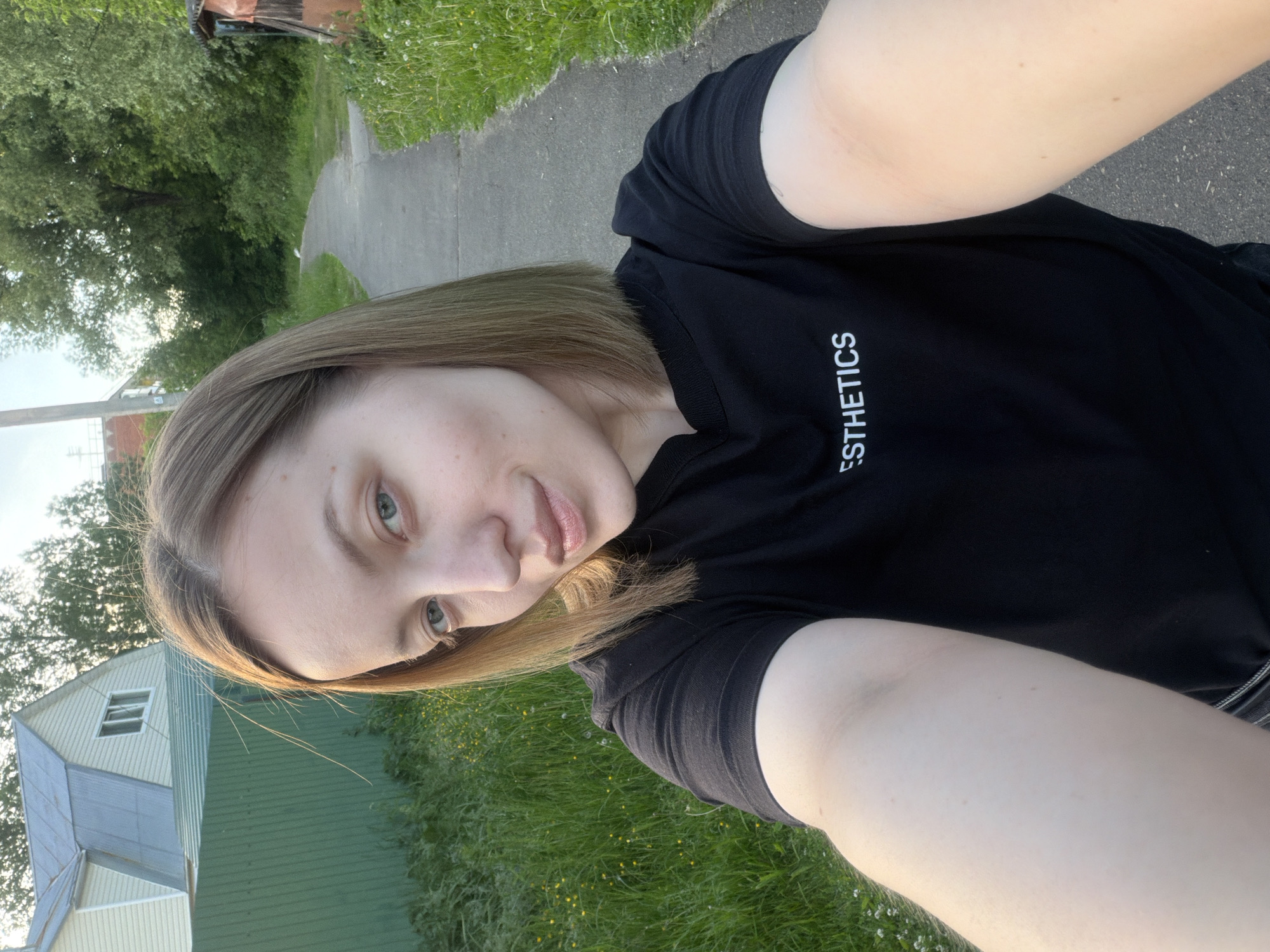 The image displays a head-and-shoulders selfie of a light-skinned woman taken outdoors in Reshetnikovo, Russia. The woman, likely in her 20s or early 30s, has medium-length, straight, light brown hair parted on the side, framing her face. She has light blue or green eyes and a subtle smile. She is wearing a black t-shirt with the white text "ESTHETICS" visible on the chest. Her right arm is extended, holding the camera or phone to capture the selfie.
The background reveals a semi-rural or residential environment. To the left, a light-colored two-story house with a multi-pitched roof is partially visible above a tall, dark green corrugated metal fence. Below the fence and house, green grass with scattered small yellow wildflowers grows. To the right and receding into the distance is a paved narrow path, likely asphalt or concrete, bordered by lush green grass and dense trees on both sides. A small, rustic-looking shed or outbuilding with reddish-brown siding is partially visible on the far right edge of the frame. The lighting suggests late afternoon or early evening, with a warm glow on the right side of the woman's face and arm. No other individuals or interactions are depicted.