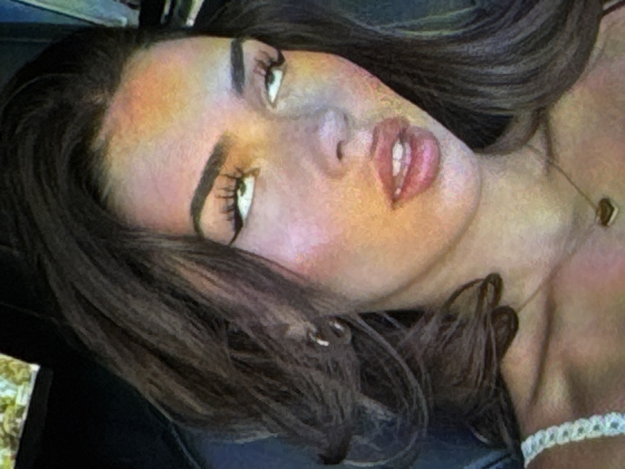 A close-up, head-and-shoulders shot of a young woman with dark, wavy hair framing her face. She is wearing a white lace top and a delicate gold necklace with a heart-shaped pendant. Her eyes are looking upwards and to the side, with dramatic winged eyeliner. The lighting casts a warm, colorful glow on her skin, with hints of orange and pink. She is wearing hoop earrings in her left ear. The background is dark and out of focus, with a hint of a screen or object in the upper left corner.