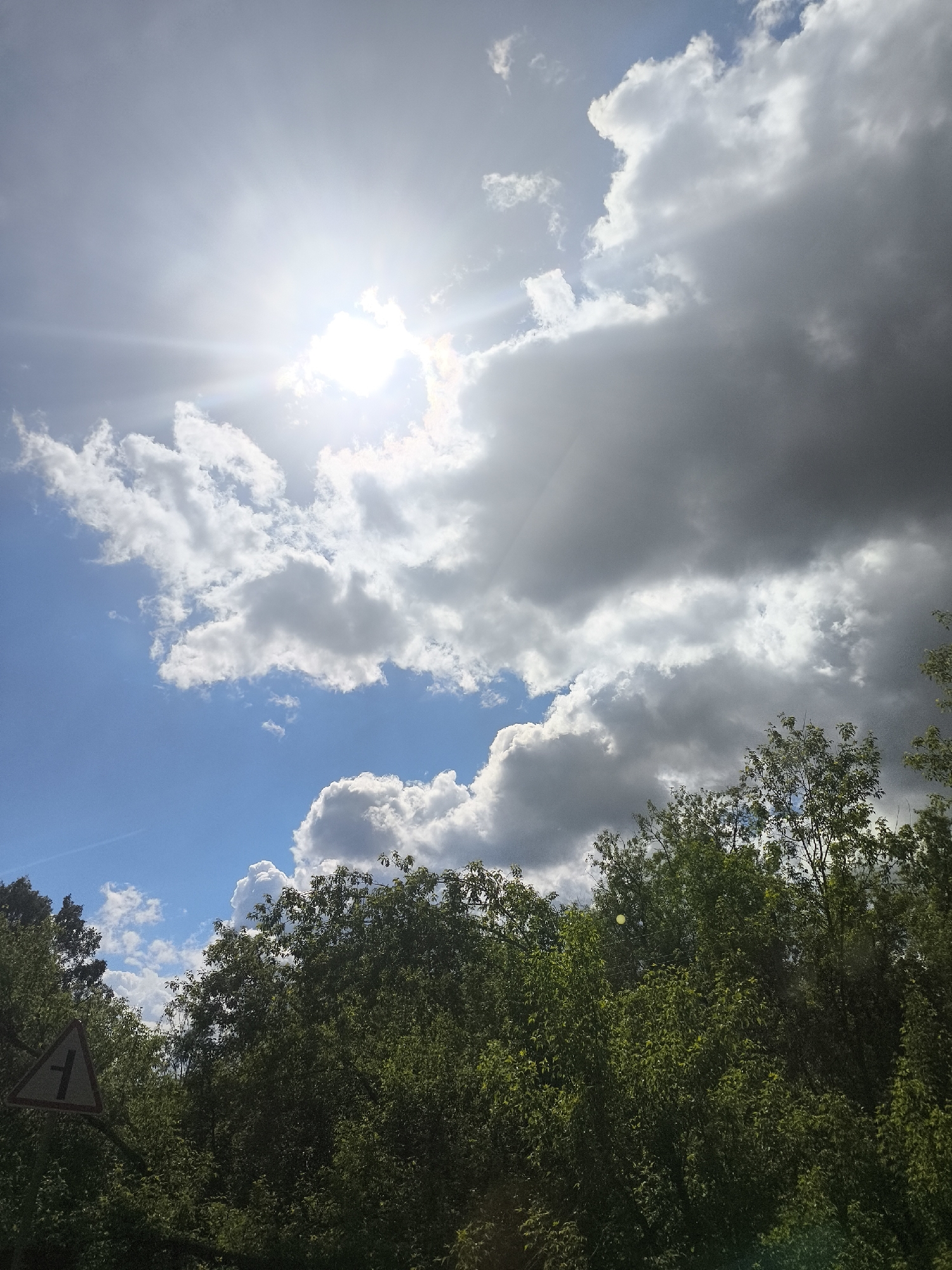 The image captures a view from a low angle, looking up towards the sky and trees. The sky is predominantly blue with a mix of white and gray clouds, and the sun is visible, partially obscured by the clouds, creating a bright glare. The trees are full and green, forming a dense canopy at the bottom of the frame. A triangular road sign is visible on the left side of the image, partially obscured by the foliage.