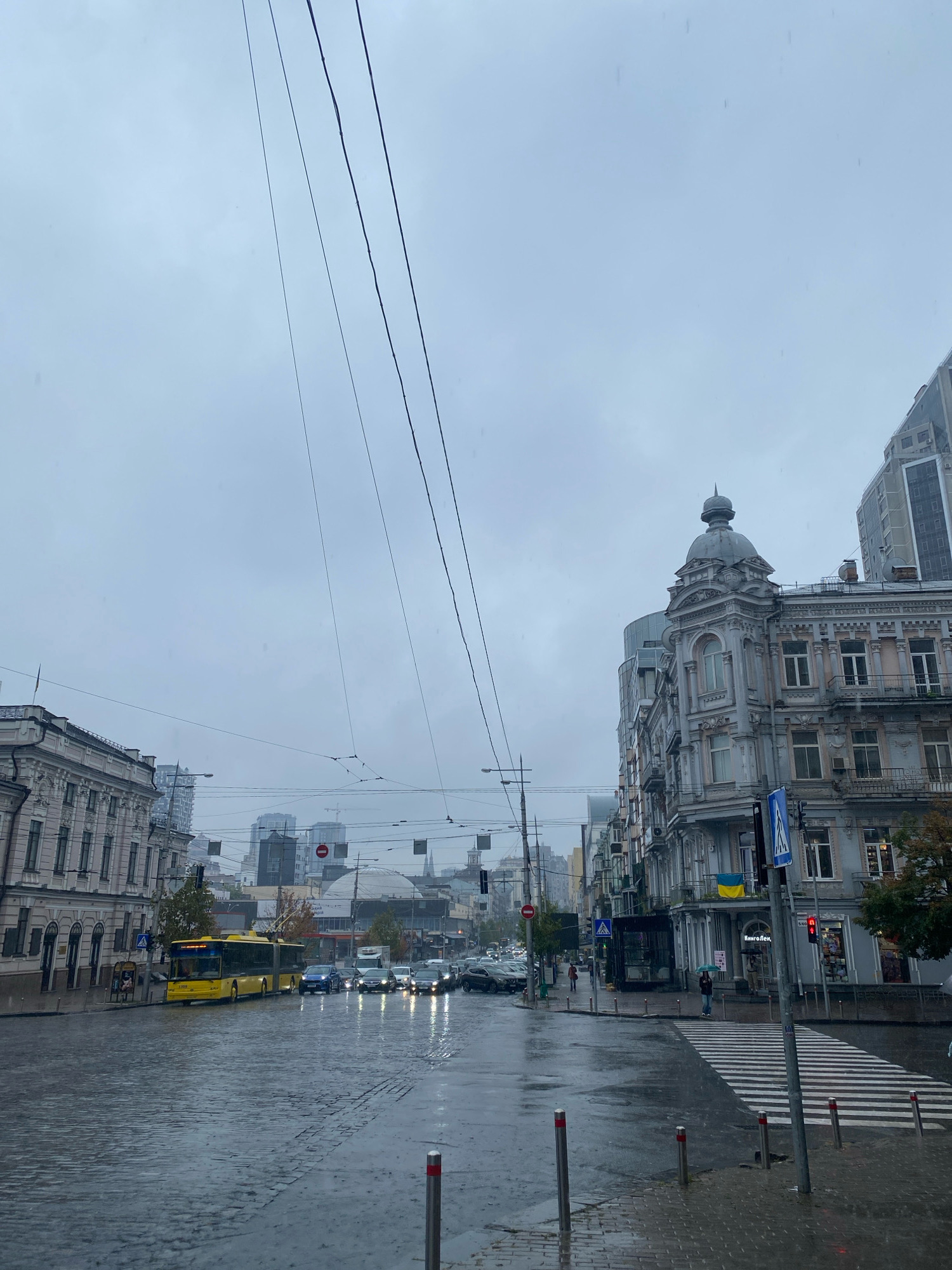 The image depicts a city street on a cloudy and rainy day. The street is wet and reflects the surrounding buildings and vehicles. On the left side of the street, there is a light-colored building with ornate architectural details. Across from it, on the right side, is a grey building with similar historical architecture, topped with a dome. A modern glass-fronted building is partially visible behind the older structures.
Several vehicles are present on the street, including a long yellow bus, several cars, and what appears to be a trolleybus. The traffic lights are visible, with one showing red. Pedestrians are present, some holding umbrellas, as they navigate the wet conditions. A pedestrian crossing is clearly marked with white stripes. Overhead wires, likely for electric transport, crisscross the sky. The overall impression is of a bustling urban environment experiencing inclement weather.