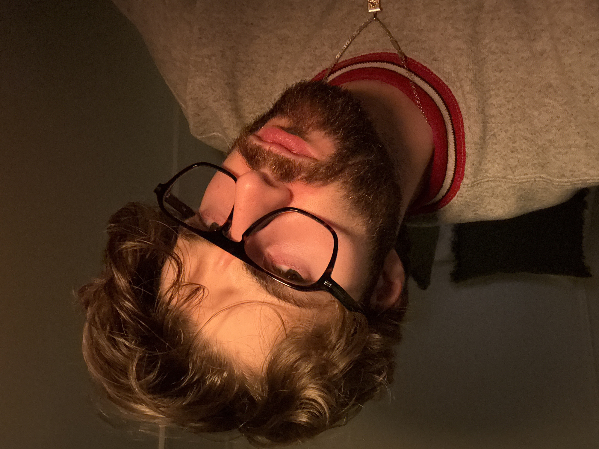 A close-up, inverted portrait of a Caucasian male with fair skin, brown wavy hair, a brown beard, and eyeglasses. The man is wearing a light gray sweatshirt with a red and white striped collar and a silver necklace with a pendant. The background is a dark, solid color. The location cannot be determined from the image.