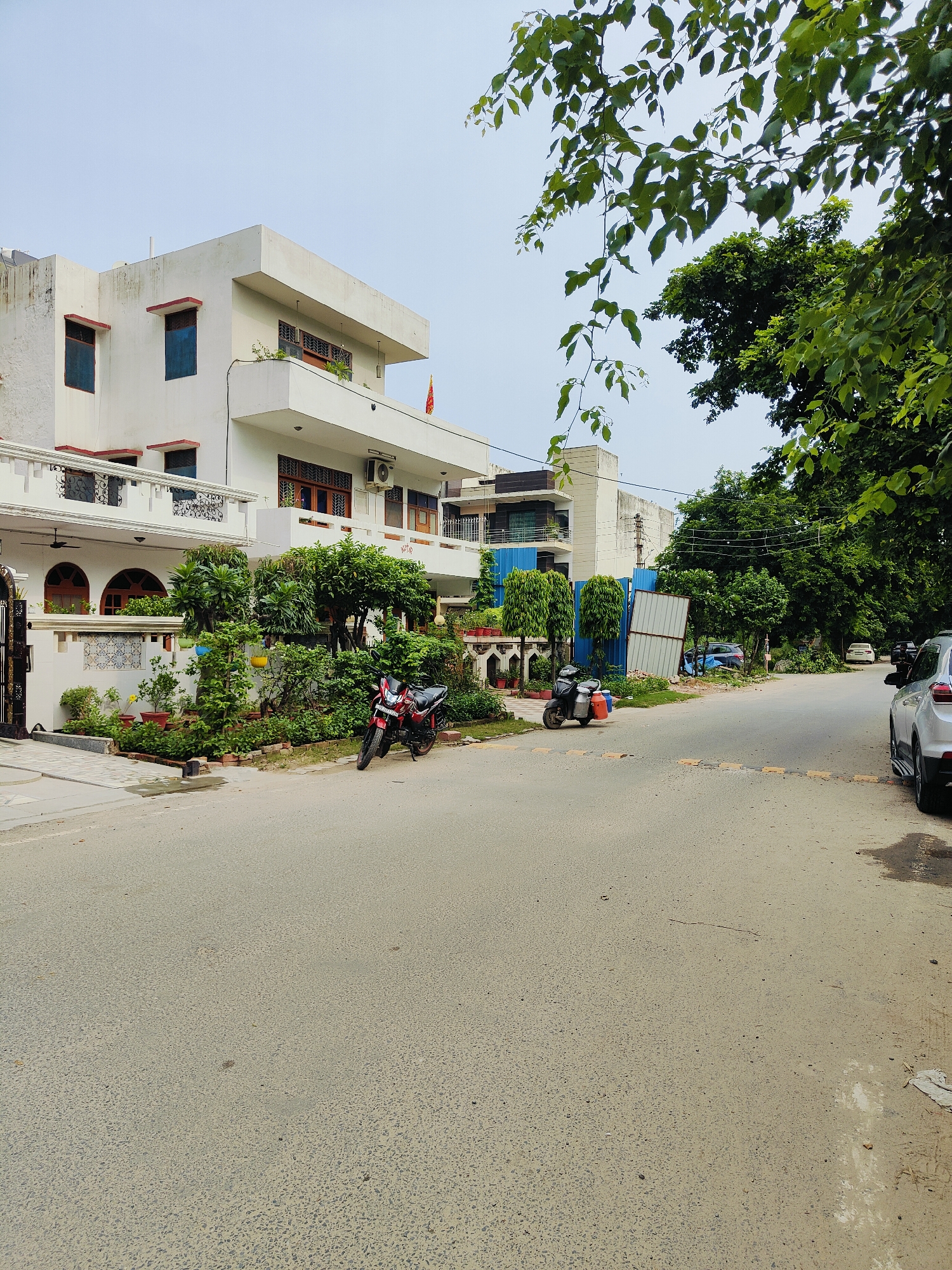 This is a street-level, eye-level, medium shot of a residential street in Gurgaon, India. The scene is dominated by a white, multi-story house on the left, with a balcony, several windows with red frames, and an air conditioning unit visible. The house has a well-maintained garden in front, with various plants and potted flowers. A red motorcycle is parked on the left side of the street, facing the viewer. To the right of the house, the street continues, with a dark gray scooter with orange saddlebags parked near a construction barrier. Further down the street, several parked cars are visible, including a silver SUV on the far right. Lush green trees line the street, and the sky is overcast. The road surface is asphalt with some patches and debris. The overall impression is of a quiet residential neighborhood.