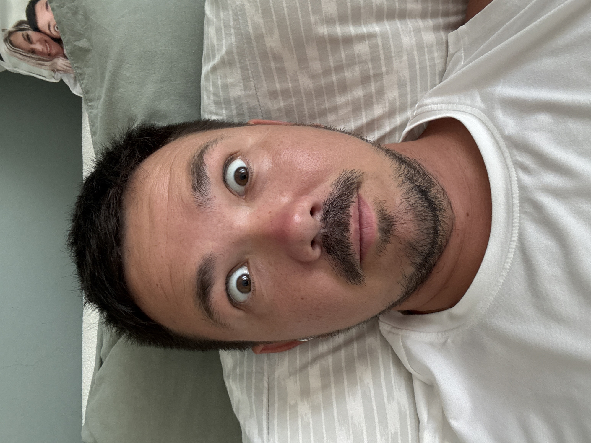 A selfie of a man with dark hair, a mustache, and a goatee, lying down on a striped pillow. He is wearing a white t-shirt and has a surprised expression with his eyes wide open. In the background, a cutout of a woman and a man is visible behind a green pillow. The environment appears to be a bedroom. The man's facial features and attire are clearly visible. The cutout figures in the background are partially obscured. The lighting suggests it is daytime. There is no discernible connection to Paphos Municipality, Cyprus.